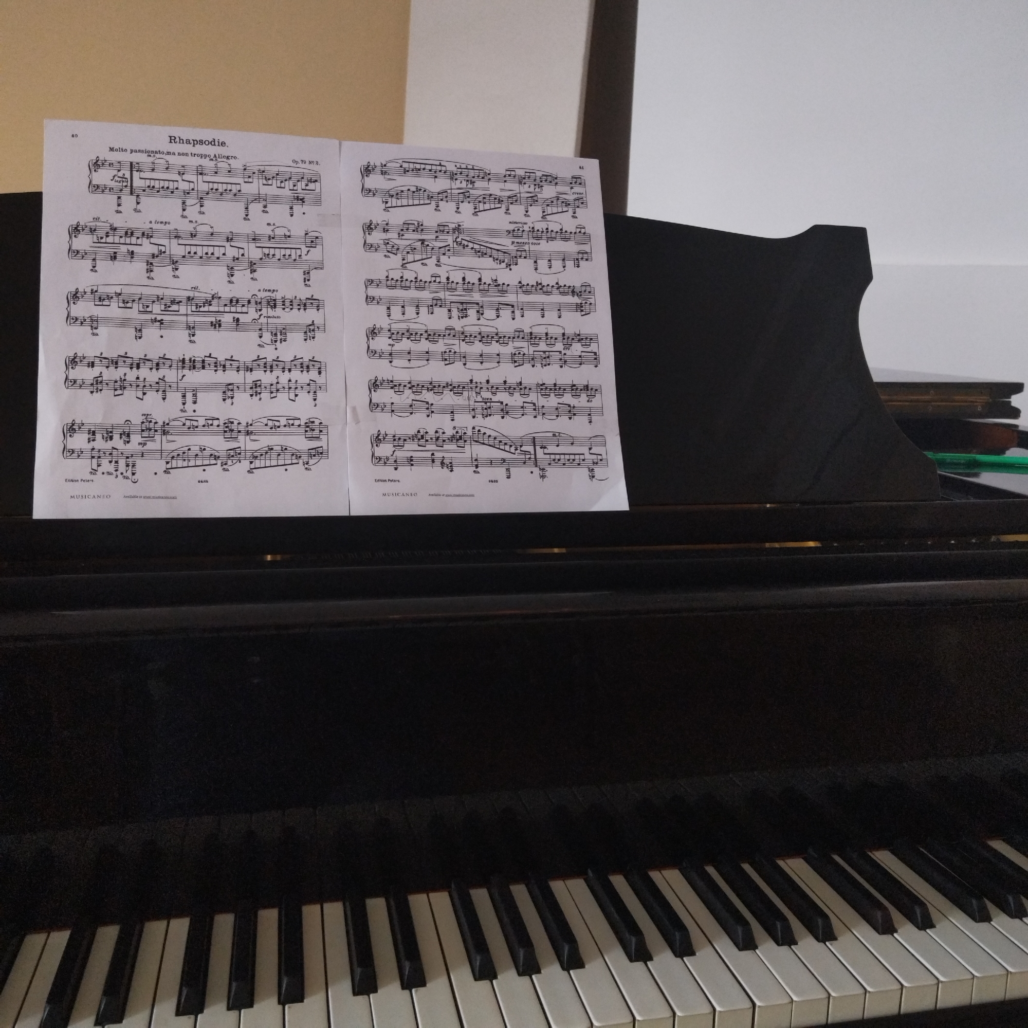 The image displays a close-up view of a piano, likely a grand or upright, with sheet music prominently placed on its music stand. The piano itself is dark, appearing black, with a clear view of its black and white keys in the foreground.
Two sheets of musical notation are open on the music stand. The left page is numbered "40" at the top left and bears the title "Rhapsodie." centered. Below the title, the tempo marking "Molto passionato, ma non troppo Allegro." is visible, along with the opus number "Op. 79 Nº 2." on the right. Further down on this page, the text "Edition Peters" and "MUSICANEO" can be read. The right page is numbered "41" at the top right and also shows "Edition Peters" and "MUSICANEO" at the bottom, along with the number "9686". Various musical dynamics and instructions such as "cresc." (crescendo) and "p mezzo-voce" are discernible on the music.
Behind the right sheet of music, a dark, rectangular object, possibly a book or case, and a thin, bright green object, perhaps a pen or highlighter, are subtly visible. The background consists of a plain light-colored wall, transitioning from a beige or cream tone on the left to a brighter white on the right, suggesting an indoor setting with ambient lighting. The piano's dark finish contrasts with the light background and the white keys. The image captures a moment where the piano is set up for playing, with the sheet music ready.
