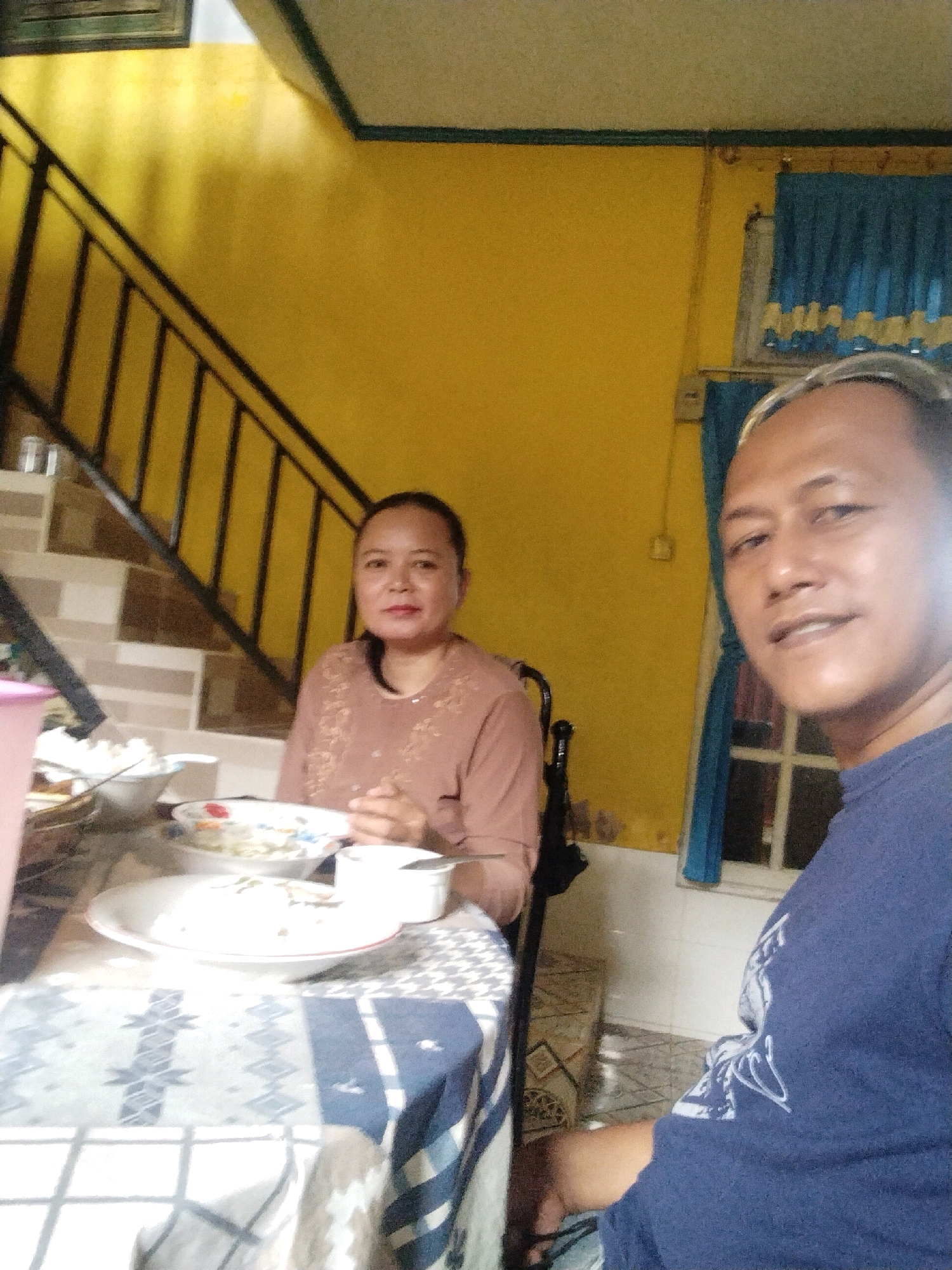 The image depicts an indoor scene in what appears to be a home in the city of Arang Limbung, Indonesia. Two individuals, a woman and a man, are seated at a table, seemingly in the process of a meal. The woman, positioned on the left side of the frame, has dark hair pulled back and is wearing a long-sleeved, light brown top. She is looking towards the camera with a neutral expression. The man, on the right side, is closer to the camera and is wearing a dark blue t-shirt. He is smiling and looking directly at the camera.
The table is covered with a patterned tablecloth, predominantly blue and white, with various dishes and bowls of food arranged on it. There is a large white bowl, likely containing rice, and other plates with food items, some of which appear to be partially consumed. A pink plastic container is also visible on the left.
The background features a bright yellow wall. To the left, a staircase with black railings is visible, suggesting a multi-story dwelling. On the right side of the image, blue curtains are drawn across a window, behind which a white wall and a grid-like structure, possibly a window pane, can be discerned. The overall environment appears to be modest and lived-in. The lighting suggests artificial indoor illumination, possibly supplemented by natural light from the window.