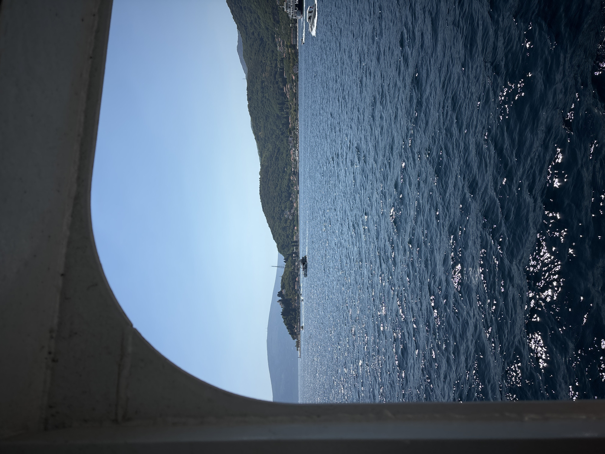 This is a photograph taken from a boat, looking out through an arched opening towards a body of water. The water is a deep blue, with sunlight glinting off the surface, creating a shimmering effect. In the distance, there is a coastline with rolling green hills covered in trees. On the hills, there are some buildings, including what appears to be a small settlement. Several boats are visible on the water, including a sailboat, a speed boat, and a larger ferry or ship. The sky is a clear, bright blue. The image captures a serene, sunny day on the water, likely in a coastal area. The presence of the boats suggests a location where water transportation is common, such as a bay or a strait. The overall impression is one of natural beauty and tranquility.
