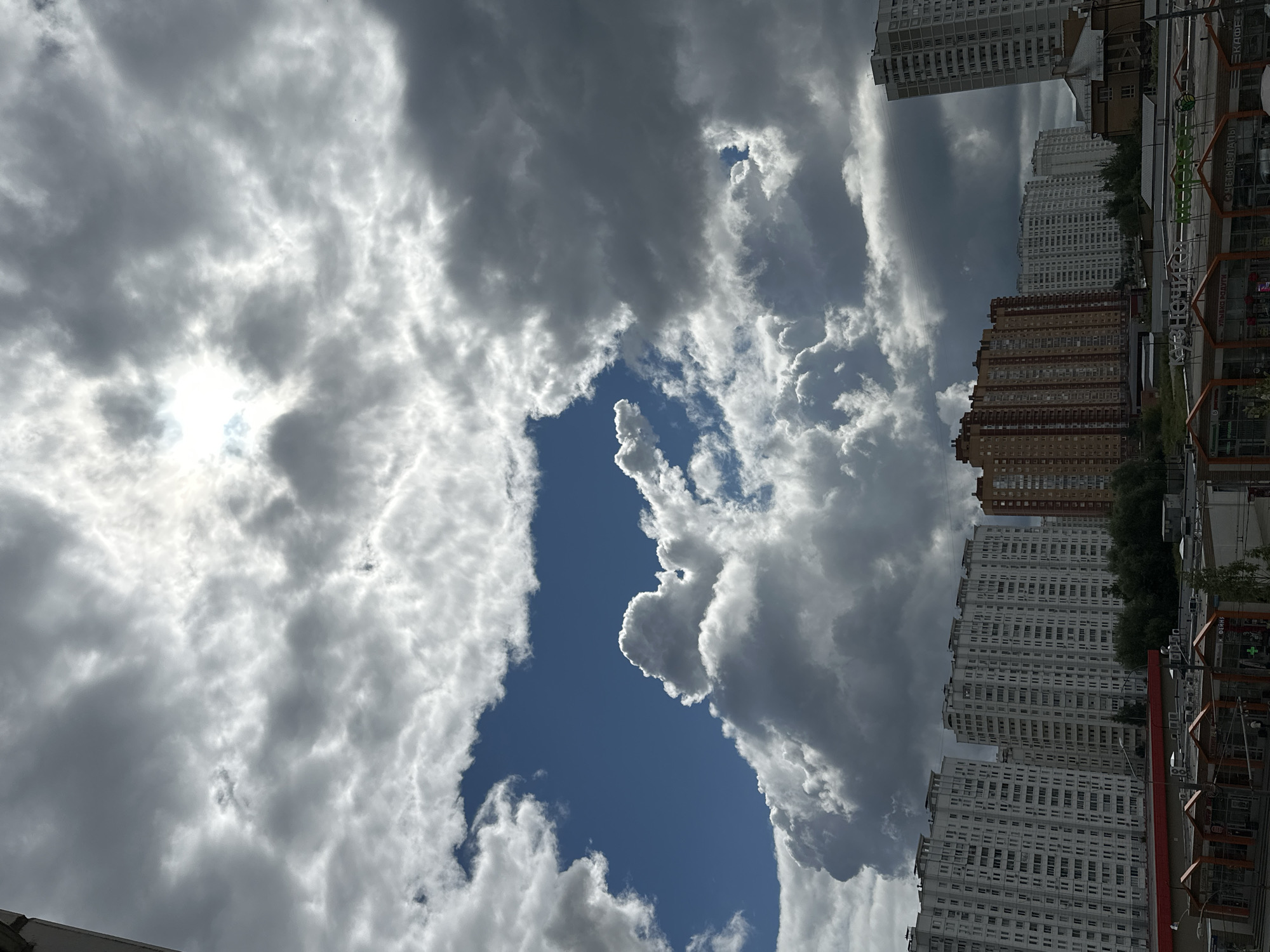 The image captures a wide sky filled with dramatic, dark, and light grey clouds, with a bright sun piercing through the clouds in the upper center. Below the sky, a city landscape is visible. Several tall, multi-story residential buildings dominate the scene, some white and others orange-brown. In the foreground, lower-level commercial buildings with storefronts are visible, some with signs in Russian. Trees and greenery are interspersed among the buildings. The overall impression is an urban environment under a dynamic, cloudy sky. The location is identified as Podolsk, Russia, a city in Moscow Oblast.
