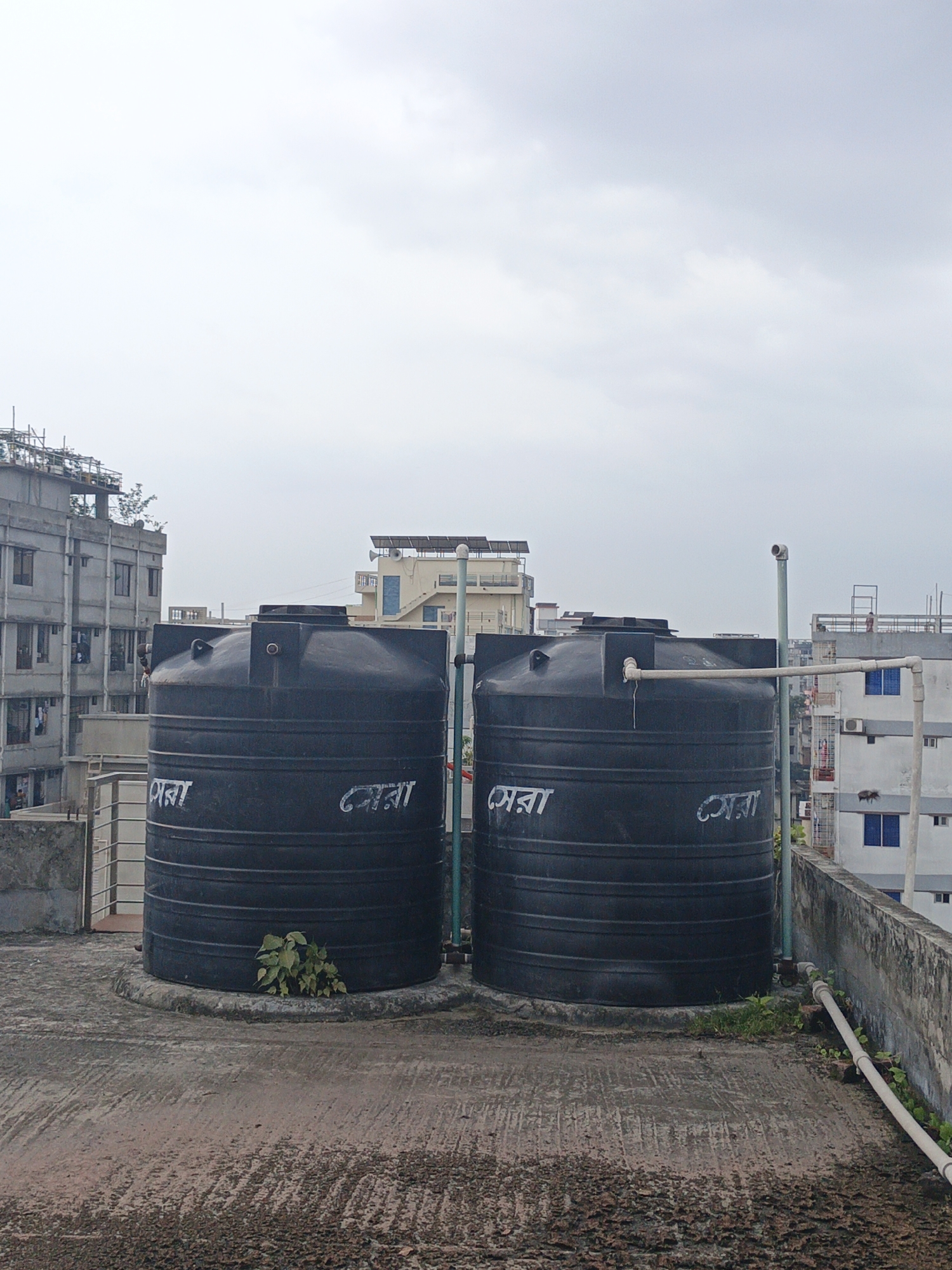 Two large black water tanks sit on a weathered rooftop in Unknown, Unknown. Buildings of varying heights are visible in the background under an overcast sky. One building has visible construction on the roof. The water tanks have white writing on them, likely brand or company name. Various pipes connect the tanks to the building. Some weeds are growing around the base of the tanks. The rooftop is worn and stained.