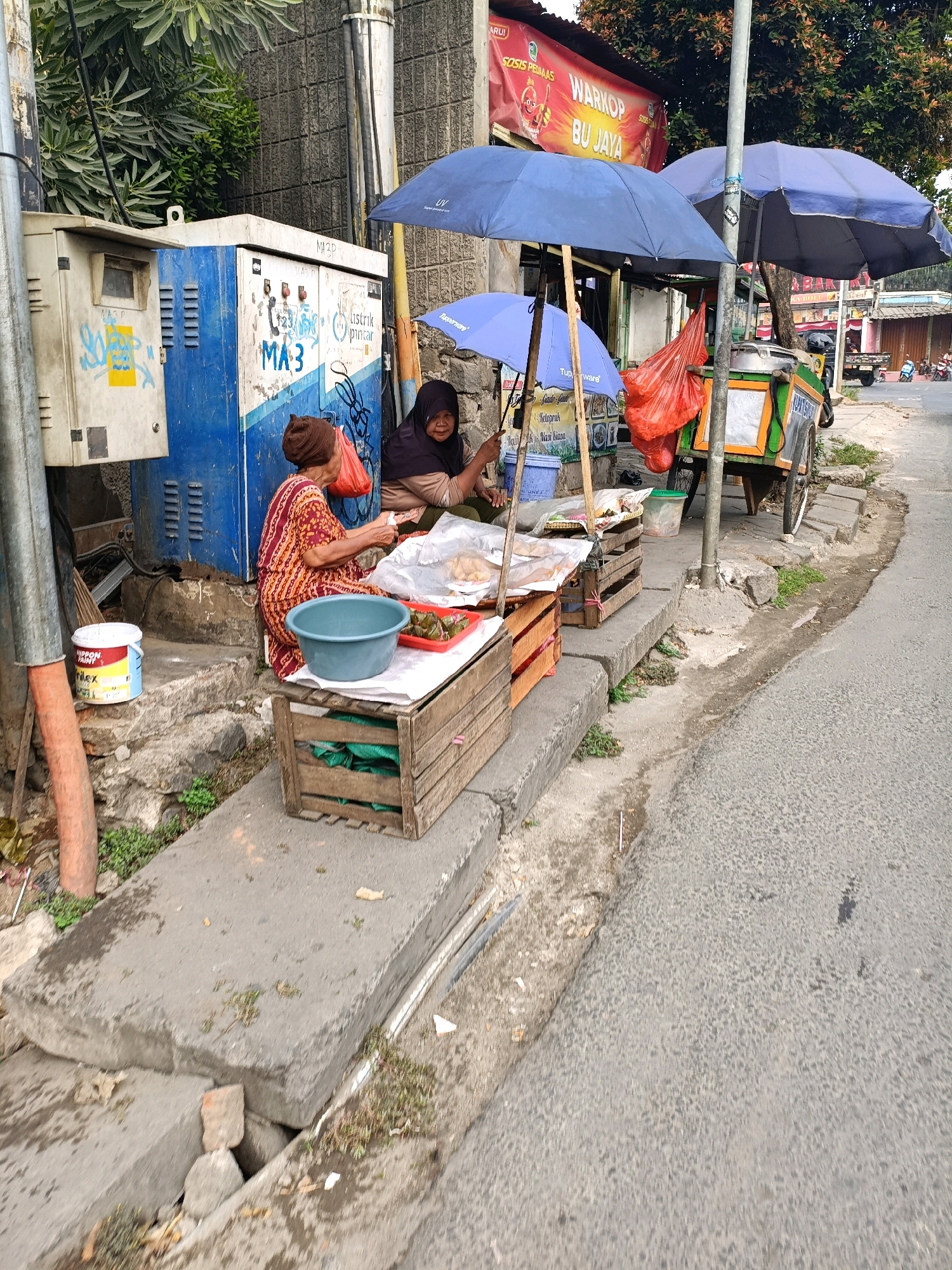 A street scene in Tangerang, Indonesia shows two women selling food from a cart and a small stall set up on the sidewalk. The women are under blue umbrellas to shield them from the sun. One woman is wearing a patterned orange and red dress and the other is wearing a dark headscarf and top. They are surrounded by various items, including baskets of food, plastic bags, and a large blue bucket. A utility box with graffiti is visible behind the women. The street is paved and there is a drainage ditch running alongside it. A sign advertising "Warkop Bu Jaya" (Bu Jaya Coffee Shop) is visible in the background. The overall impression is of a bustling street market, typical of urban Indonesia.