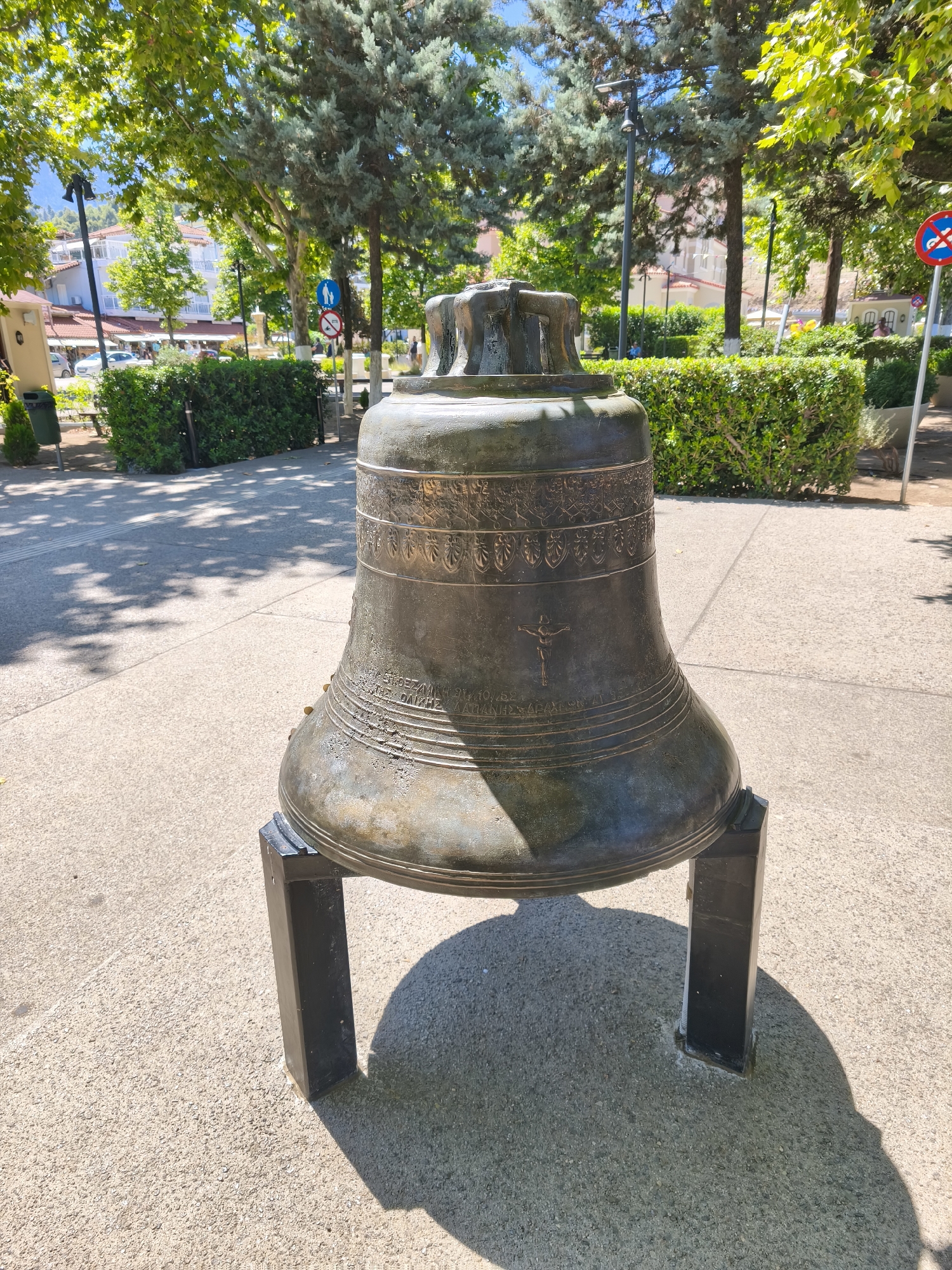 A large, weathered bronze bell sits on a concrete surface in what appears to be a public square or park in Prokopi, Greece. The bell is supported by two black, rectangular stands. It is decorated with bands of relief carvings, including what looks like a cross with a figure of Jesus Christ. The background shows lush green trees, manicured hedges, buildings with white facades, and a clear blue sky. Sunlight casts shadows on the ground, indicating a bright, sunny day. The area appears to be well-maintained and accessible to the public.