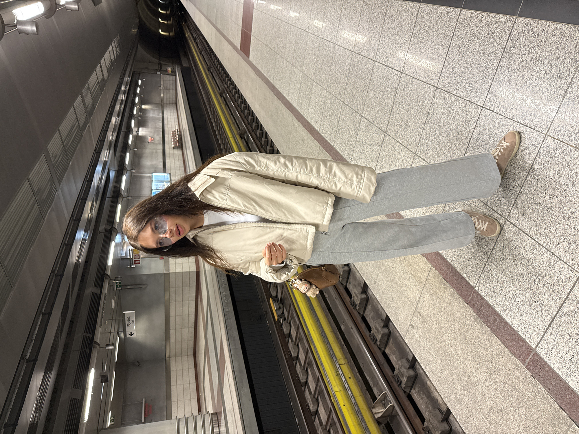 A person stands in a metro station. The person is wearing light grey sweatpants, a white shirt, a tan bomber jacket, and tan shoes. The person is also wearing sunglasses. The station has grey and brown tiled floors and a dark ceiling with lights. The train tracks are visible.