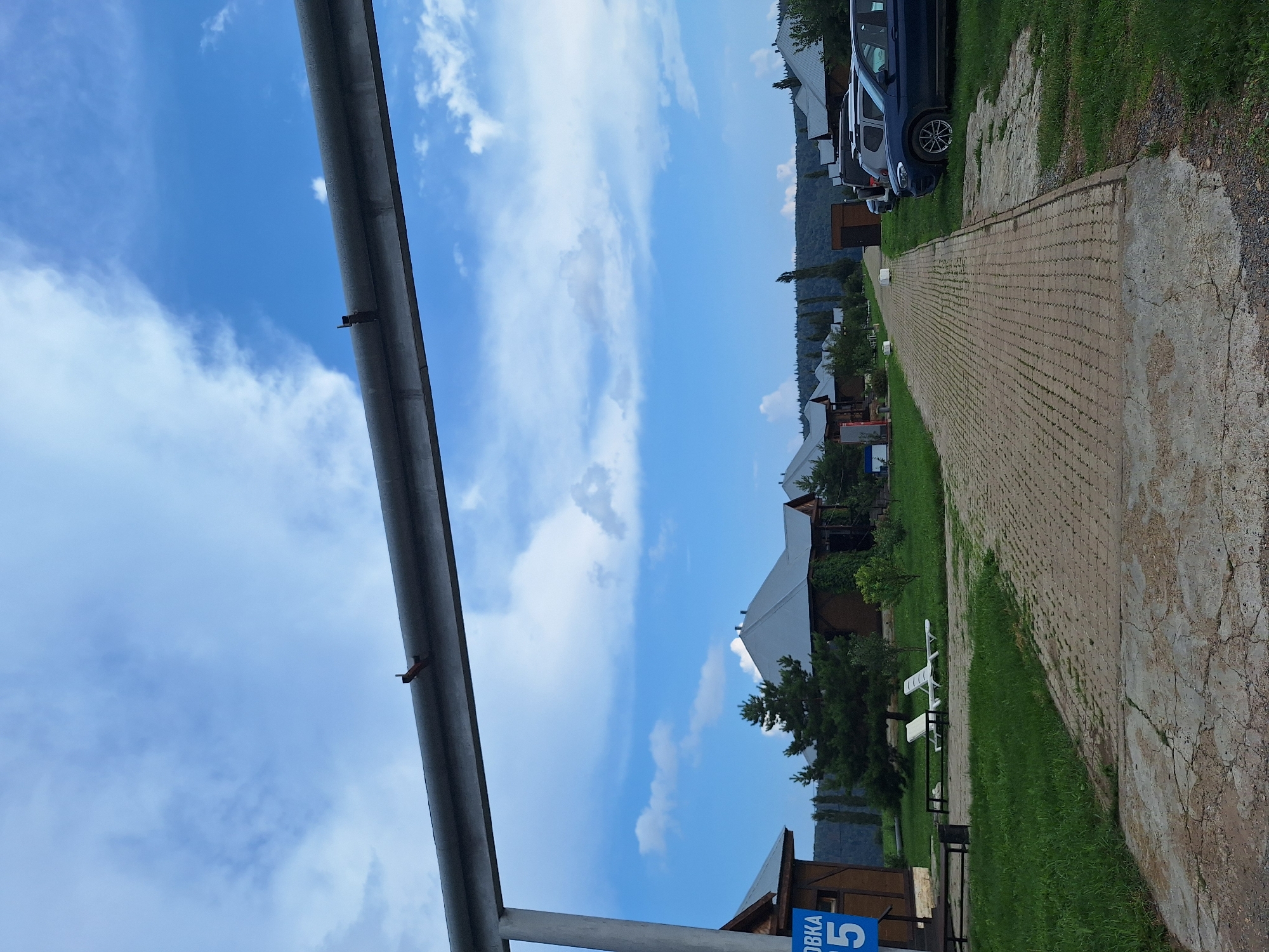 The image depicts a paved pathway leading through a village-like settlement with wooden houses featuring gray roofs. The pathway is lined with green grass and scattered trees. On the left side of the path, there are two white lounge chairs on the grass, and a blue sign with white Cyrillic lettering and the number "5". Several cars are parked on the right side of the path. In the background, a dense forest covers the rolling hills under a blue sky with scattered white clouds. A large metal pipe runs horizontally across the upper portion of the image, supported by vertical structures. The overall scene suggests a peaceful, rural setting. The location is identified as Atamanovka, Russia.