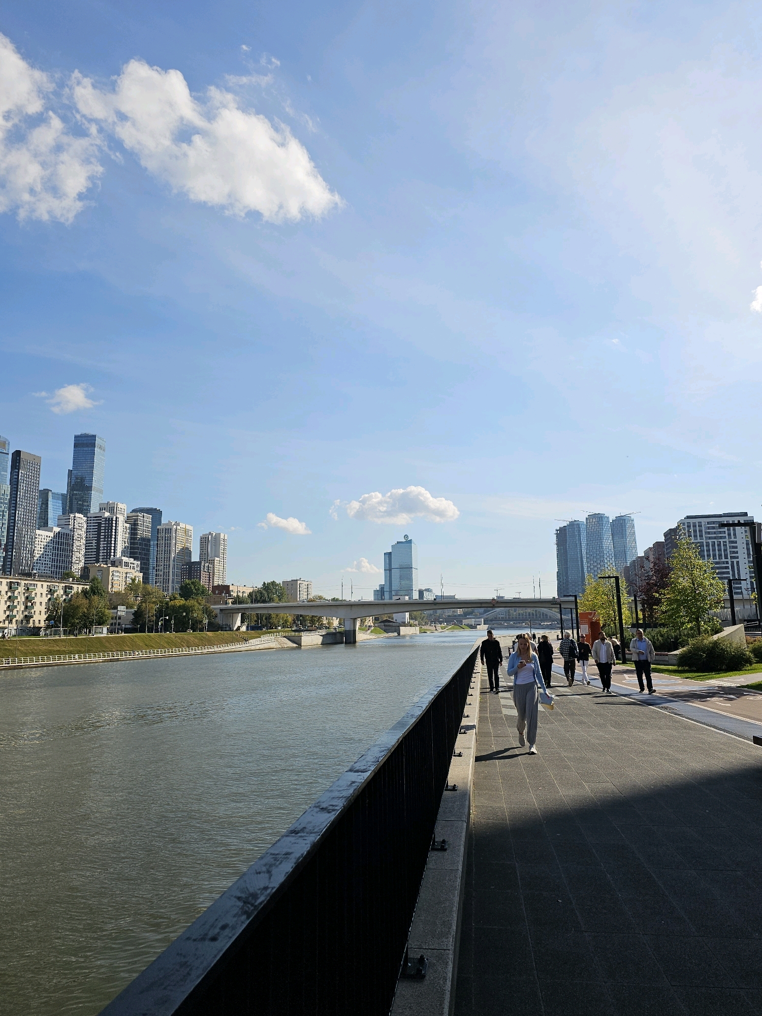 The image captures a sunny day in Moscow, Russia, featuring a view of the Moscow River and a modern cityscape. In the foreground, a paved walkway runs alongside the river, bordered by a dark metal railing. Several people are walking along the path, some individually and some in small groups. The river is calm, reflecting the blue sky and white clouds. Across the river, the Moscow skyline is dominated by a cluster of contemporary skyscrapers, including the prominent triangular building of the Neva Towers and the white cylindrical building of the Dominion Tower. A bridge spans the river in the mid-ground. The overall atmosphere is one of urban activity and leisure on a pleasant day.