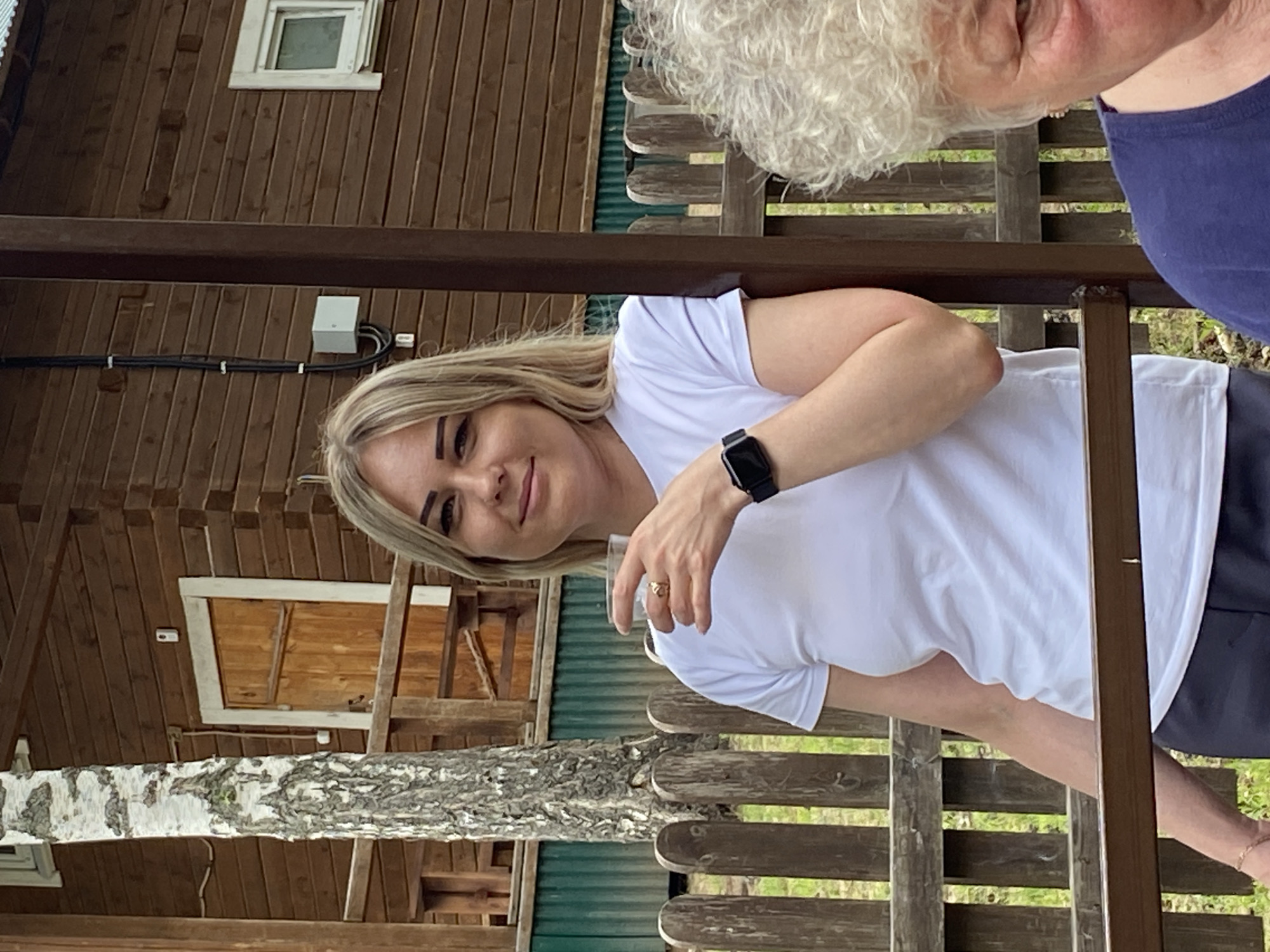 The image shows two people, a woman and an older person, in an outdoor setting with wooden structures. The woman in the foreground has blonde hair and is wearing a white t-shirt and a black smartwatch. She is holding a clear glass and appears to be smiling. There is a ring on her finger. The older person, partially visible on the right, has curly white hair and is wearing a dark blue tank top. They are both seated on a wooden bench or seating area, with a wooden fence behind them and a wooden building with a window in the background. A birch tree trunk is also visible in the foreground. The setting appears to be rustic, possibly a dacha or a rural retreat in Noviy, Russia. The lighting suggests it is daytime, and the overall atmosphere seems relaxed and casual. There is no visible text in the image.