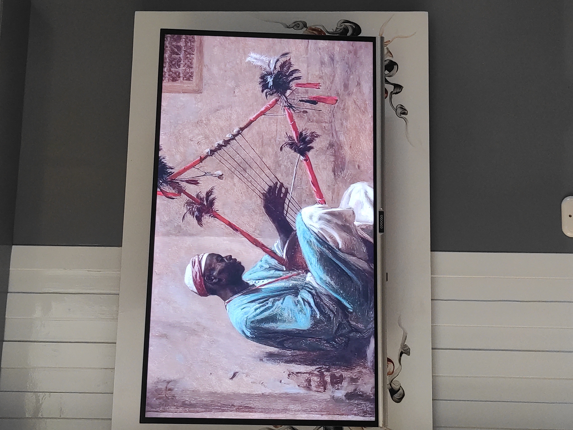 A digital painting of a dark-skinned man playing a stringed instrument is displayed on a television screen. The man is wearing a teal robe and a red-and-white headscarf. He is seated and facing to the right, with his hands on the instrument, which is triangular and decorated with feathers and red ribbons. The background of the painting is a muted, textured wall with a small window. The television is mounted on a wall that has vertical white panels and grey sections. Decorative, dark, swirling designs are present on the white frame surrounding the television screen. The overall setting appears to be indoors, possibly a living room or common area. The image does not contain any information indicating it is located in Sumedang, Indonesia.
