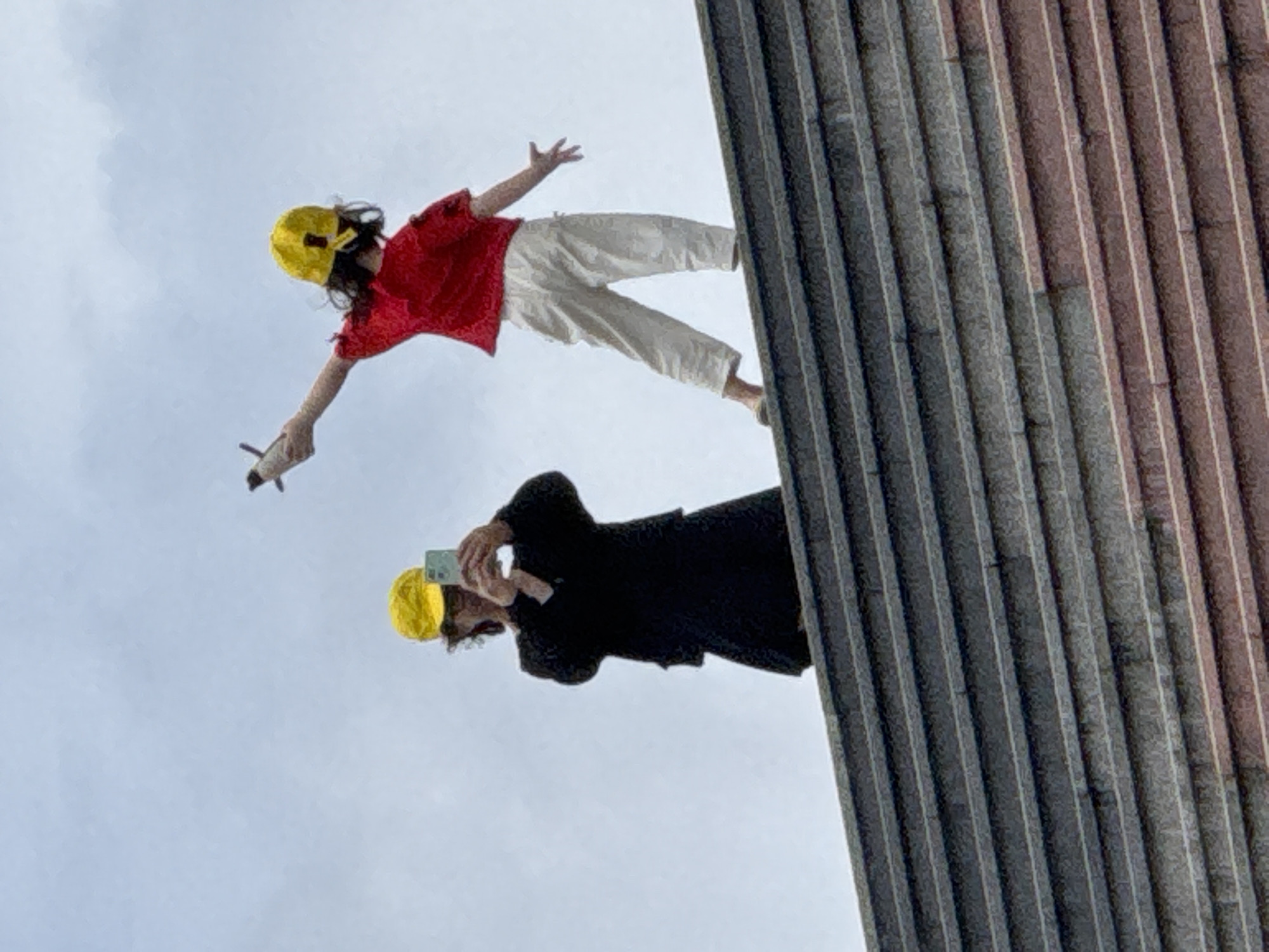 Two people, both wearing yellow baseball caps, stand on a wide, tiered staircase. On the left, a person in a black outfit is holding up a smartphone, appearing to take a photo or video. On the right, a child in a red shirt and white pants is holding a toy plane in their outstretched right hand, with their left arm extended upwards, palm open. The background consists of an overcast sky. The tiered staircase is made of dark grey stone with lighter grey or white edging on each step. This scene is located on the outdoor steps of the Da Lat Flower Garden in Da Lat, Vietnam.