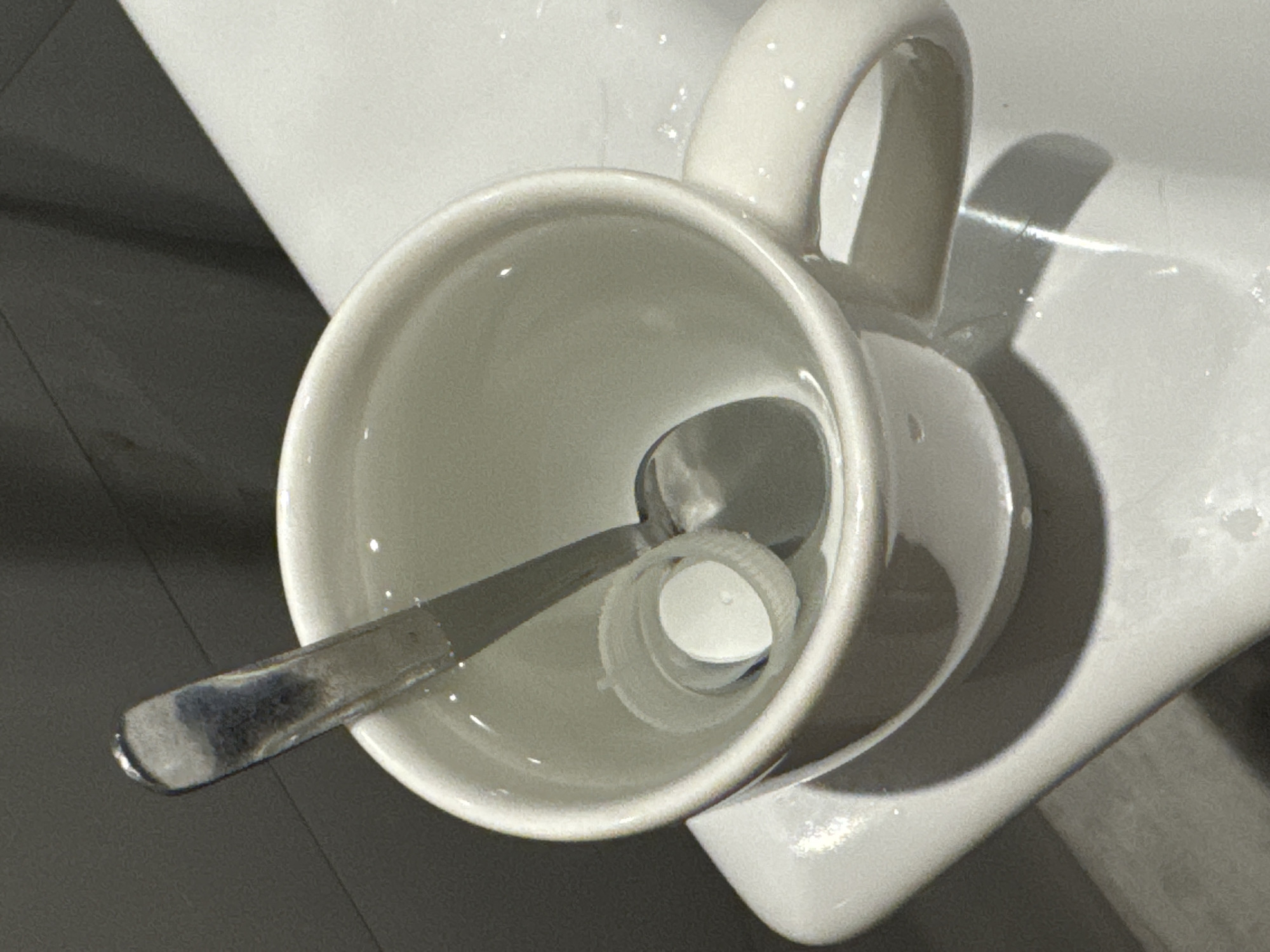 An overhead, eye-level shot shows a white ceramic mug filled with clear liquid. A silver spoon rests inside the mug, with its handle extending out of the liquid. A small, white plastic cap is submerged in the liquid, positioned beneath the spoon. The mug is on a white surface, likely a sink or countertop, with a dark, textured wall in the background. The lighting is dim, casting shadows on the scene. There are no people present in the image. The scene appears to be indoors, possibly in a bathroom or kitchen. There is no information in the image to suggest this location is in Aksu, Turkey.