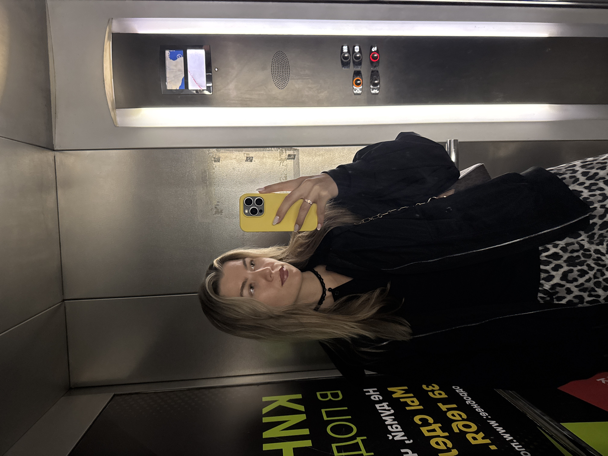 The image captures a person taking a selfie in an elevator in Unknown, Unknown. The elevator has a gray interior with a panel of floor buttons and a display. The person is holding a yellow smartphone and is wearing a black top, a choker necklace, and possibly a leopard-print skirt or pants. The elevator also contains informational signage with text in an unidentified language.