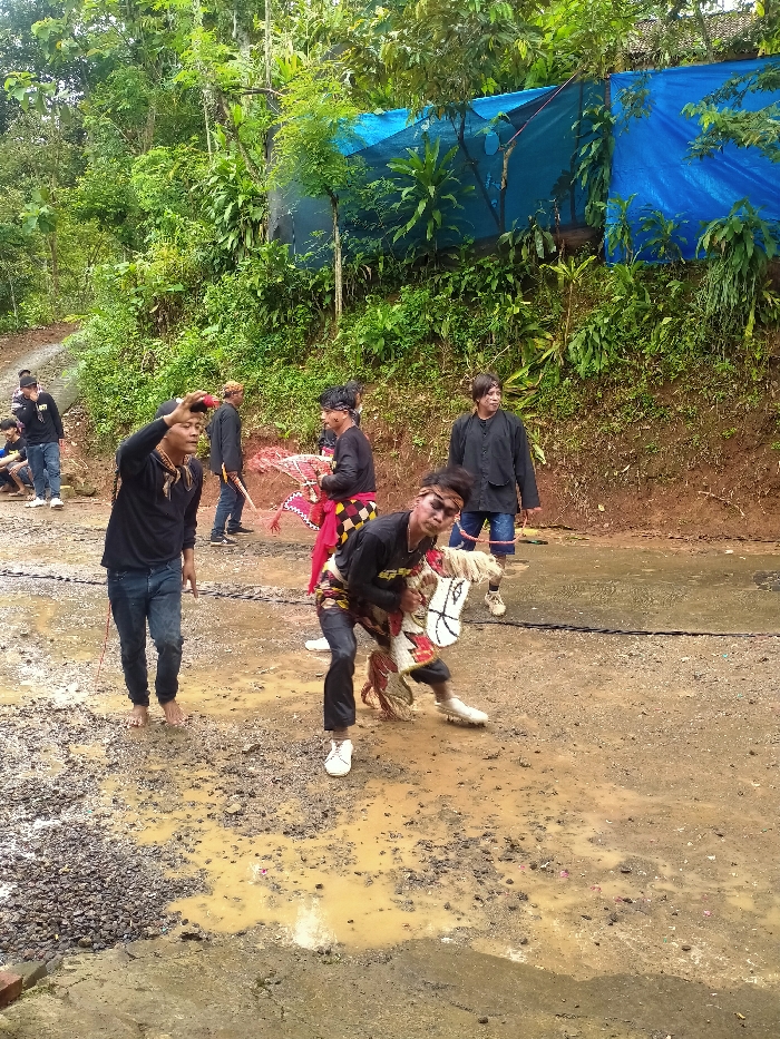 The image captures an outdoor scene in Gempolan, Indonesia, depicting a cultural performance on a muddy, gravelly, and wet dirt ground. In the central foreground, a male performer, wearing a black long-sleeved shirt, dark pants, and white shoes, is actively engaged with a white and brown Kuda Lumping (hobby horse) prop, appearing to dance or pose. He has visible face paint and a headband. To his left, another male, dressed in a black long-sleeved shirt and blue jeans, barefoot, holds his right hand up to his forehead, with a patterned scarf around his neck. In the mid-ground, other individuals are present: one male in black clothing with a red and black patterned sarong holds an ornate fan-like object, while another male in black with a traditional head covering holds a stick-like prop. Further right, a male in a black button-up shirt and dark pants stands observing. Several other figures are partially visible in the far left background, some seated. The background consists of a dense green hillside covered with lush tropical foliage and trees. A large blue tarp is stretched horizontally across the upper right section of the hillside, indicating a temporary covering or structure. The ground shows signs of recent moisture, with numerous puddles and wet patches.
