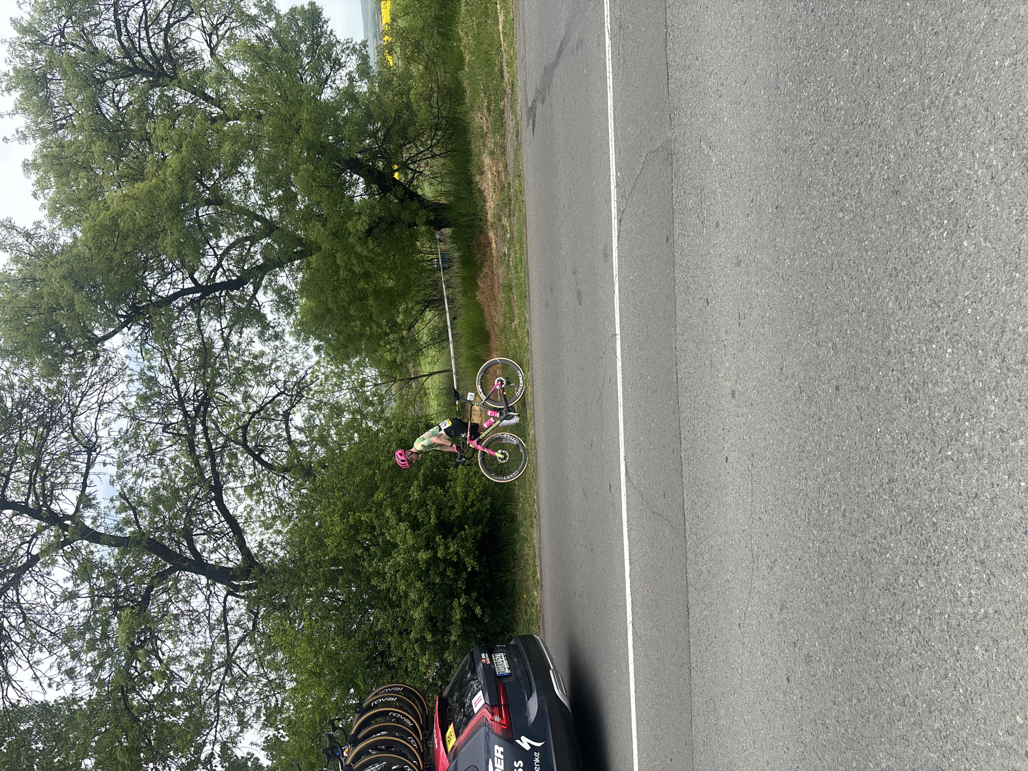 The image shows a roadside scene near Burgas, Bulgaria. A cyclist wearing a green and pink outfit and a pink helmet stands next to a pink and black bicycle on the grassy shoulder of a road. A dark-colored support vehicle is parked nearby, equipped with multiple spare bicycle wheels on its roof. The road is asphalt with a white line marking the edge. Trees and vegetation line the shoulder, and a yellow field is visible in the distance.