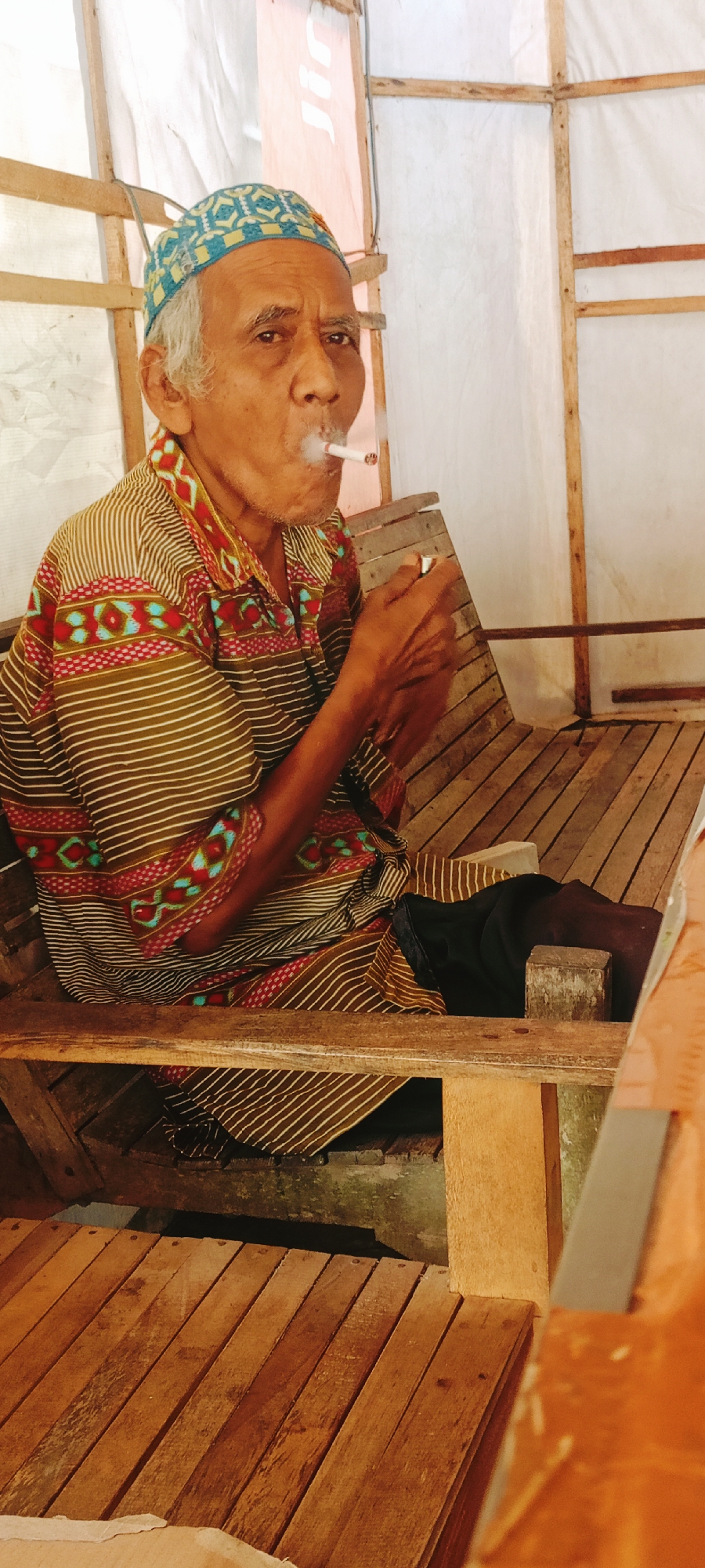 An elderly man with gray hair and a patterned cap is seated on a wooden bench, smoking a cigarette. He is wearing a striped shirt with colorful geometric patterns. His right hand is holding a lighter near the cigarette. The bench is made of rough-hewn wooden planks, and the setting appears to be an outdoor or semi-open structure with wooden supports and white sheeting. The ground beneath the bench is also made of wooden planks. The scene has a rustic, informal feel. The location is identified as Lhokseumawe, Indonesia.