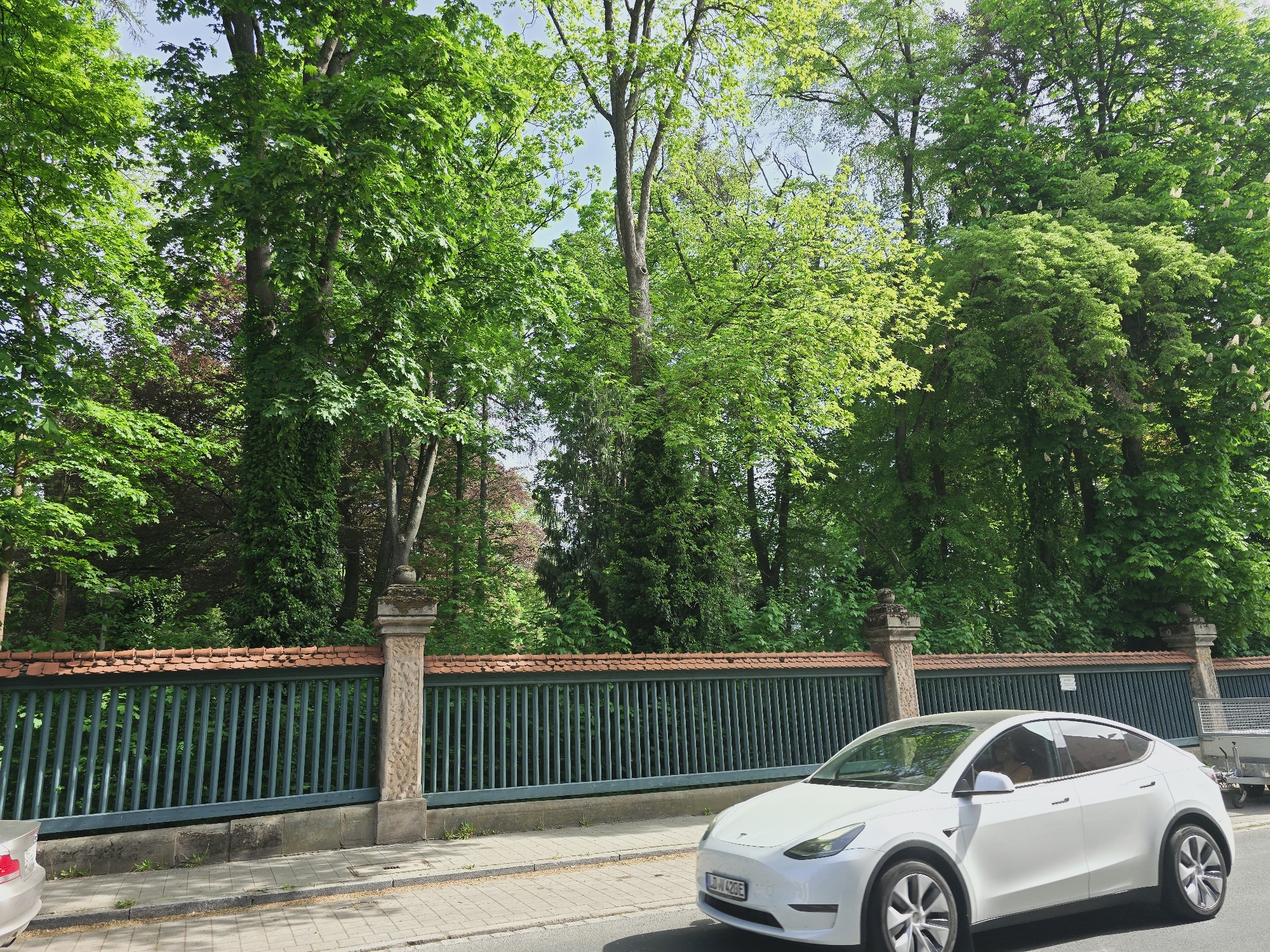 A white Tesla Model Y is parked on the street in Zirndorf, Germany. The car is next to a long, dark green fence with ornate stone pillars. The fence has a red tiled top. Lush green trees fill the background. Part of another car is visible in the bottom left corner, and a trailer is seen behind the Tesla on the right. The street is paved with grey bricks. The license plate on the Tesla is visible. The sky is visible through the trees.
