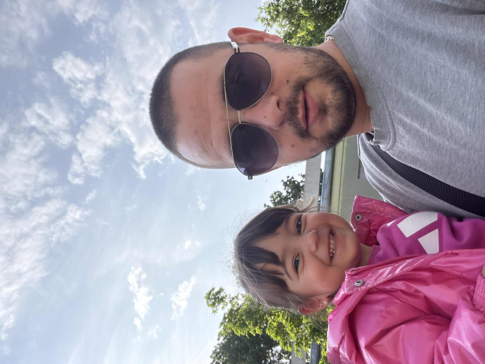 A selfie-style, outdoor portrait captures a man and a young girl. The man, positioned above and behind the girl, has a shaved head, a beard, and wears aviator-style sunglasses and a gray sweatshirt. The girl, in the foreground, smiles brightly and wears a pink, puffy jacket over a pink shirt with white accents. The background consists of a partly cloudy sky and green foliage of trees. The photograph was taken in Burgas, Bulgaria.