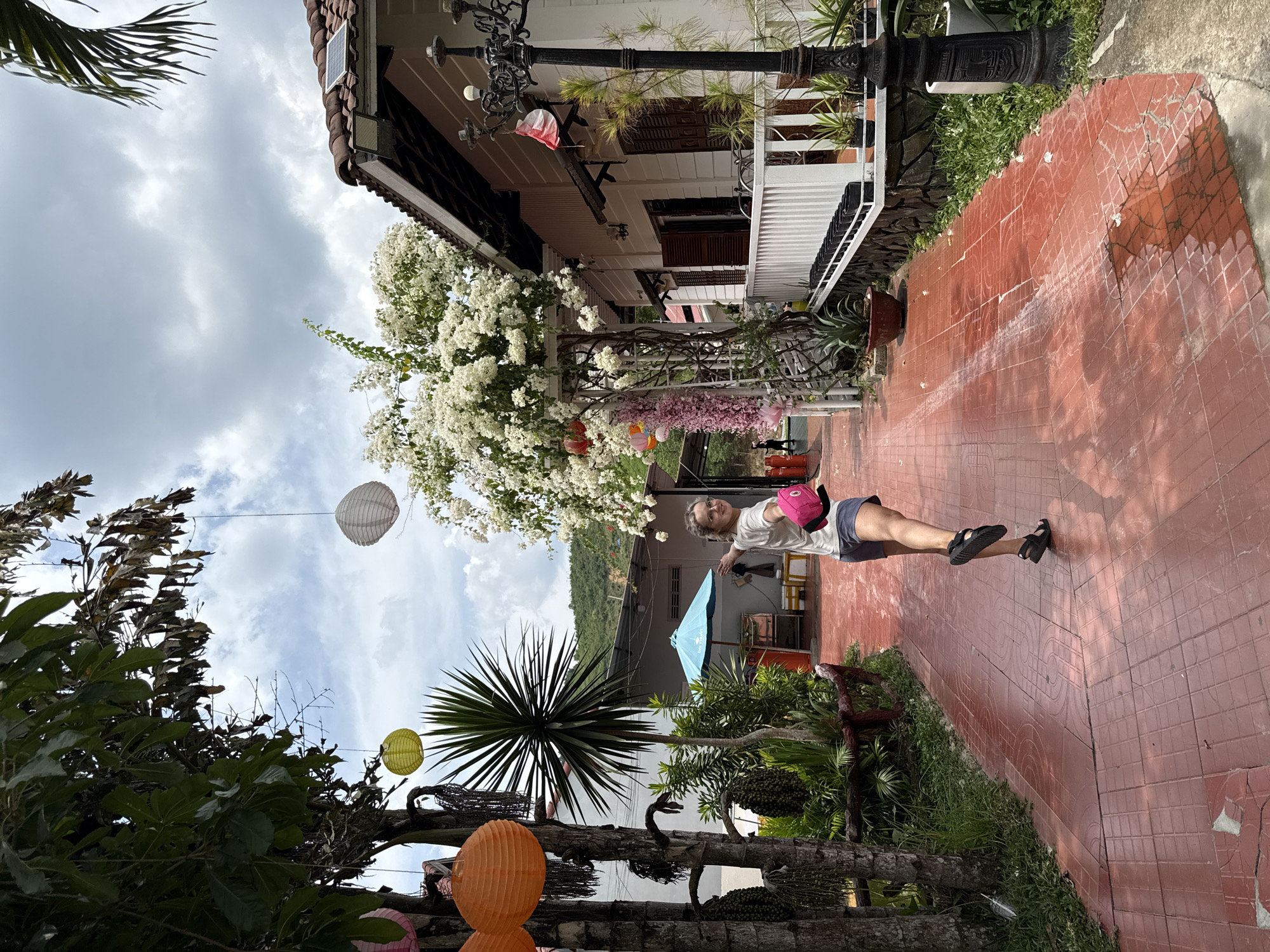 A person is standing on a red brick pathway, slightly angled towards the camera, while wearing a white t-shirt, gray shorts, and black sandals. The person is holding a pink object and facing forward. Behind them is a two-story building with white siding, brown shutters, and a red tiled roof, partially obscured by a large white flowering tree. To the left of the building, the sky is visible with scattered white clouds. Below the tree, various plants and trees surround a blue umbrella and some outdoor furniture. In the foreground, colorful paper lanterns hang from the trees. The location is Nam Khánh Vĩnh, Vietnam.