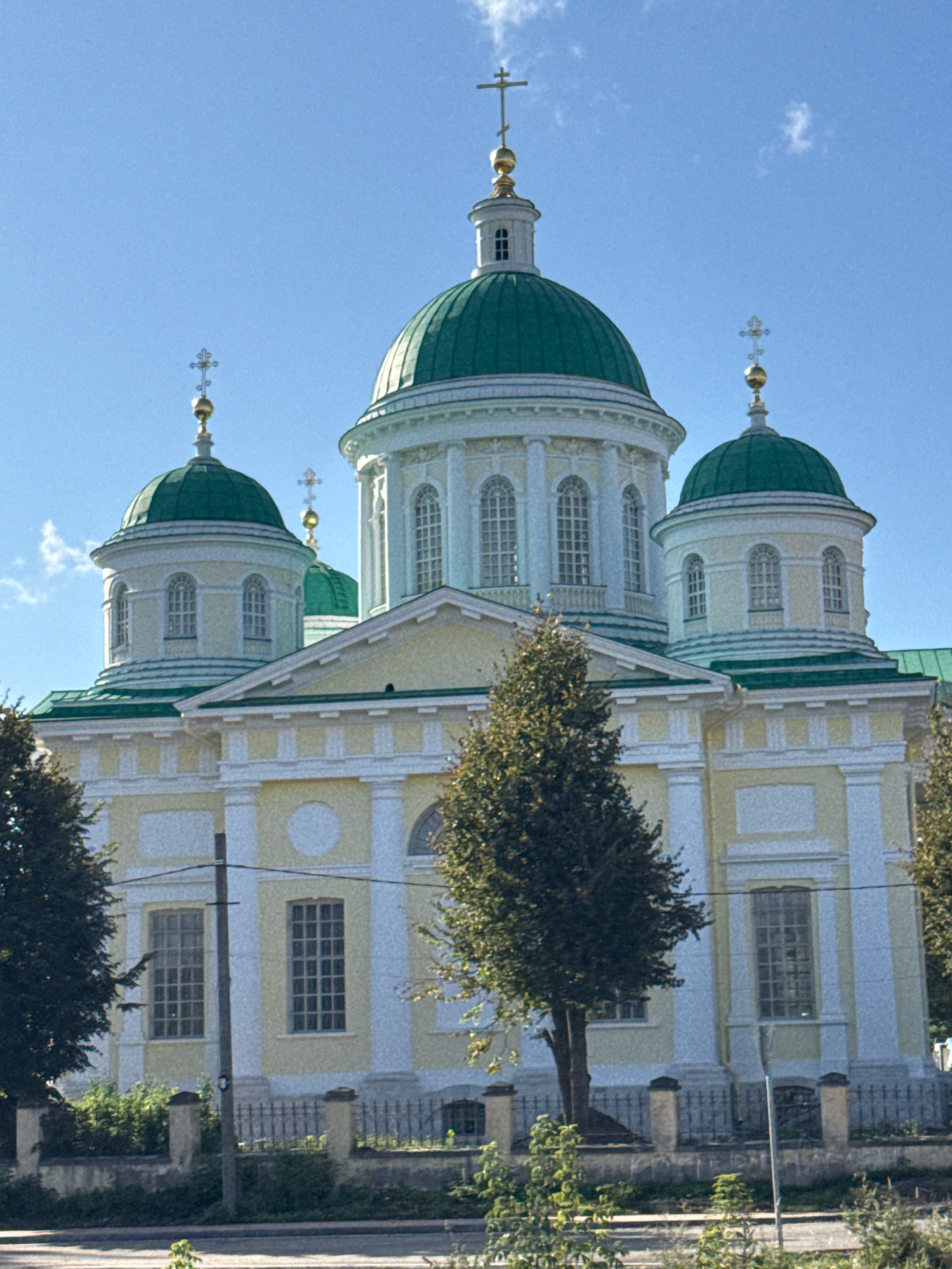 The image shows the Borisoglebsky Monastery in Torzhok, Russia, on a clear day under a bright blue sky. The monastery's main building features a large, central green dome topped with a golden cross, flanked by smaller green domes also adorned with crosses. The facade is painted in pale yellow and white, with numerous arched windows. Trees are visible in the foreground, and a fence runs along the front of the monastery grounds. The overall scene depicts a peaceful, architectural landmark.