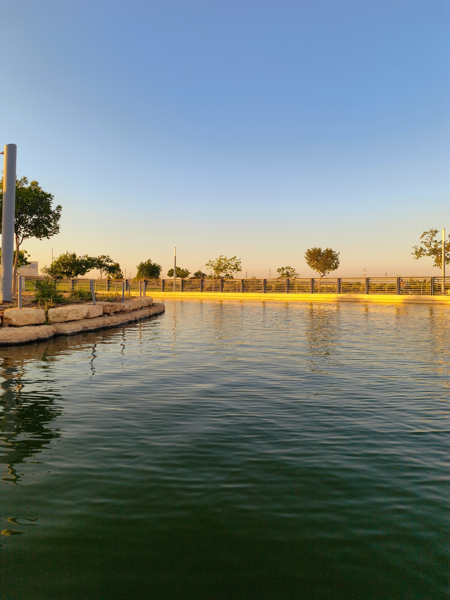 The image depicts an outdoor scene at a water feature, likely a pond or canal, in Be'er Sheva, Israel, during sunset. The sky is a clear gradient of light blue to pale yellow. On the left, a light-colored pole and some green trees are visible. The water in the foreground has a dark green hue with ripples, reflecting the sky and surrounding elements. Along the edge of the water, there is a retaining wall made of large, light-colored stones, with some greenery behind it. Beyond this, a fence with horizontal metal bars runs along the scene, illuminated by the warm, golden light of the setting sun. Trees are scattered along the fence line, their reflections also visible in the water. In the distance, a few structures and utility poles can be seen. The overall impression is one of a peaceful, urban park setting bathed in the warm glow of twilight.