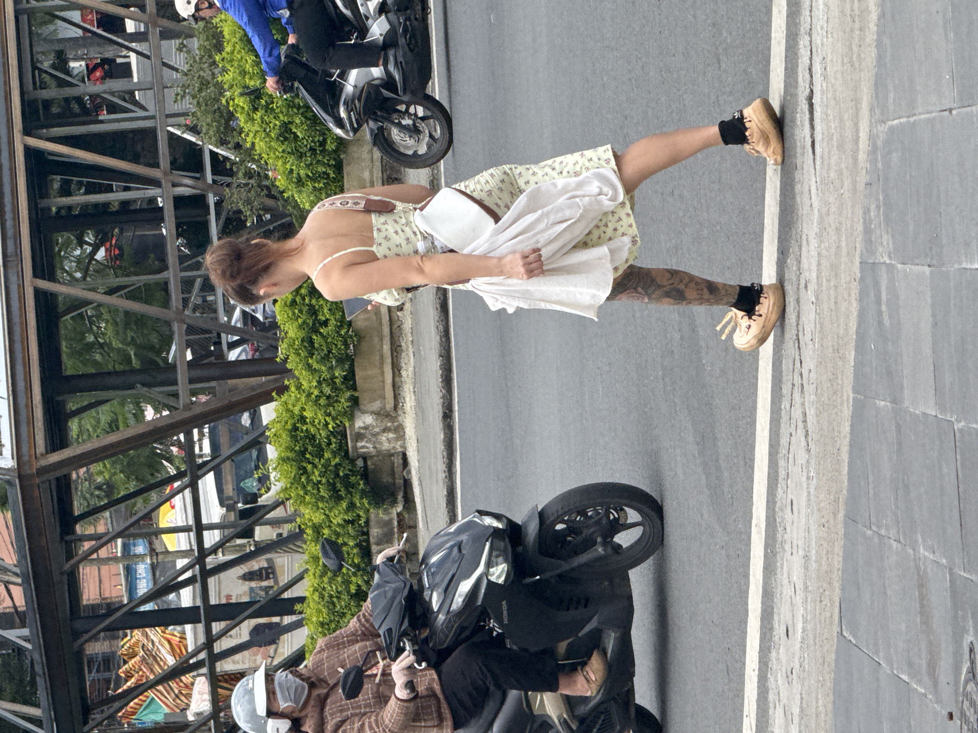 This image captures a daytime street scene in Da Lat, Vietnam. The foreground consists of a gray tiled sidewalk transitioning to an asphalt road, marked by a solid white line.
Three individuals and two scooters are visible. On the left, an individual, likely an older adult, is seated on a black Honda Airblade scooter. They wear a light gray helmet, a white surgical mask, a brown and white checkered long-sleeved jacket, and dark pants with flat brown shoes. Their hands are on the scooter's handlebars.
In the center, a younger adult female is walking across the asphalt street, moving away from the camera. Her reddish-brown hair is styled in a bun. She is dressed in a light yellow, floral-patterned spaghetti strap dress, black ankle socks, and tan, thick-soled shoes. A white cross-body bag with a patterned brown strap is worn over her right shoulder. She holds a folded white garment in her left hand and a black smartphone in her right hand, at which she is looking down. Her left leg features extensive dark tattoos, visible from mid-thigh down.
On the far right, partially visible, an adult male rides a black Honda scooter. He wears a white helmet and a blue long-sleeved shirt with dark pants. Only the front wheel and a portion of his body and the scooter are within the frame.
Behind the street and vehicles, a continuous row of green bushes extends across the mid-ground. Above the bushes, a prominent multi-level structure composed of dark metal beams, arranged diagonally and horizontally, spans the scene. Further in the background, behind this metal structure, urban elements include various buildings, trees, and a white vehicle (possibly a truck or van) with colorful materials draped over it. A partially visible blue sign displaying Vietnamese text, "ONG CHU," can also be seen. The lighting indicates daylight conditions.