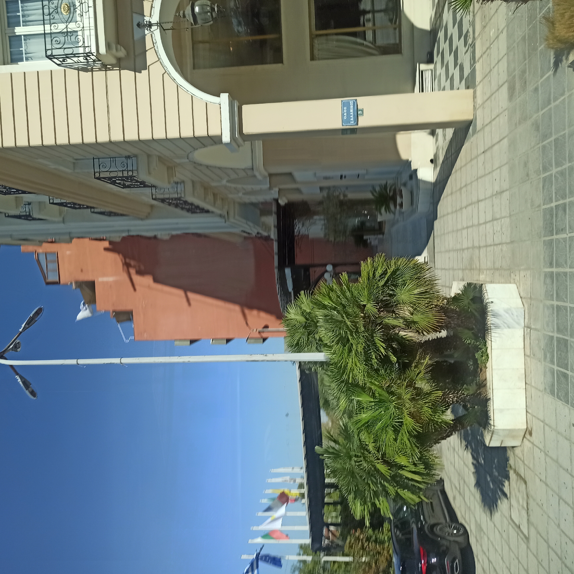 The image captures an outdoor urban scene, likely in Thessaloniki, Greece, given the presence of Greek signage and flags. In the foreground, lush palm trees are planted in white marble planters. To the left, a black SUV is parked on a light-grey tiled walkway.
Dominating the background are two buildings. The building on the right is light beige with ornate balconies featuring black wrought iron railings. The building on the left is a darker, reddish-orange color. Several flags are visible on poles in front of the reddish-orange building, including what appears to be the Greek flag. A pedestrian bridge with a dark roof spans across the scene.
A street sign on the beige building reads "ΟΔΟΣ ΣΑΛΑΜΙΝΟΣ" (Odos Salaminos), indicating Salaminos Street, and the number "3". A lamppost is visible in the upper left of the frame. The sky is a clear, bright blue. The scene appears to be an entrance to a commercial area or hotel complex.
