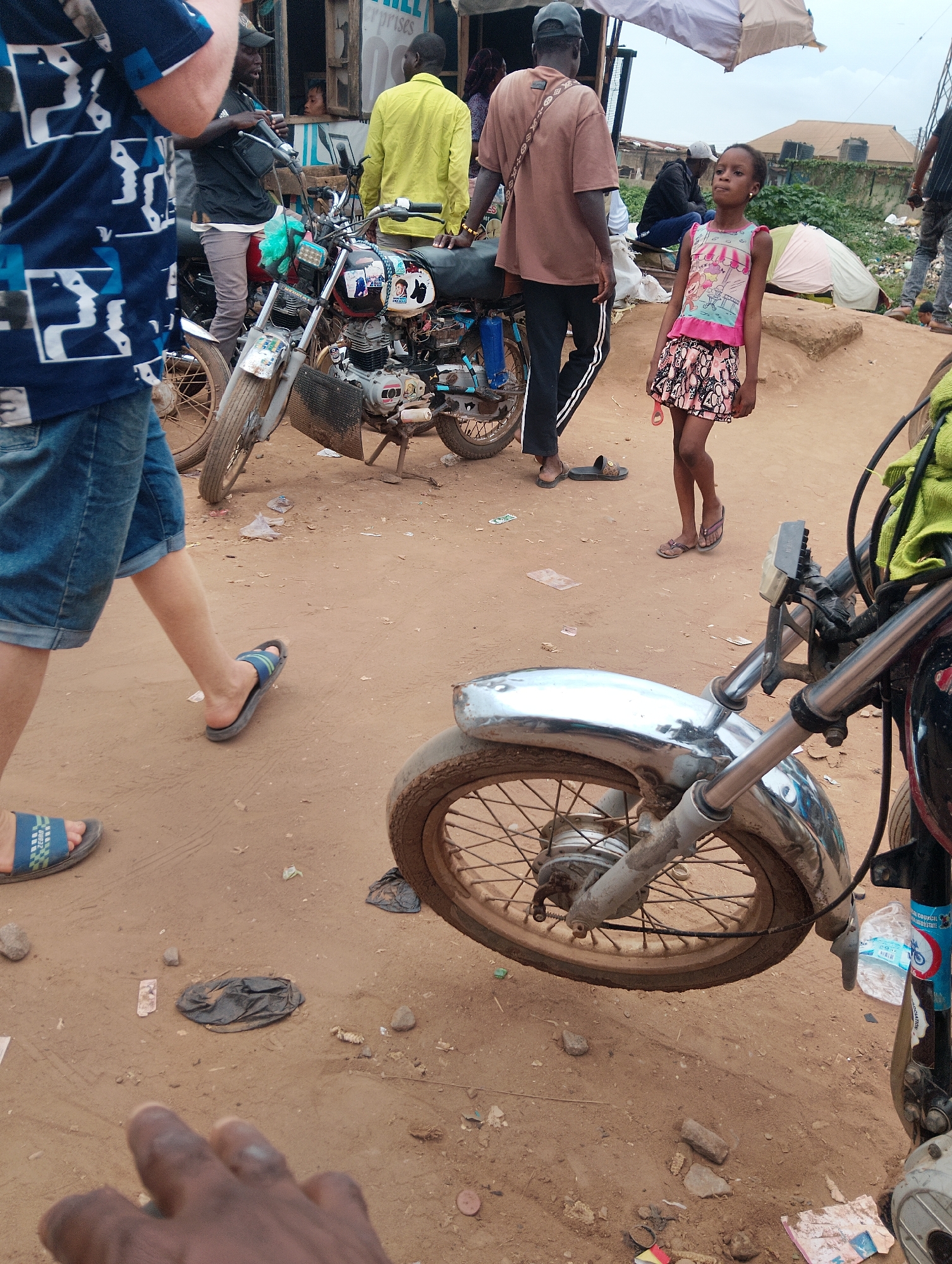 A group of people are gathered on a dirt road in Ijoko, Nigeria. In the foreground, a person wearing blue denim shorts and a blue and white patterned t-shirt walks past a parked motorcycle. The motorcycle is partially visible, with its front wheel and handlebars in the frame. Further back, a young girl in a pink and white patterned dress stands and looks upwards. Behind her, a man in a light brown t-shirt and black pants stands next to another motorcycle. Other individuals are visible in the background, some near a makeshift stall or shop with goods displayed. The environment appears to be a bustling outdoor market or street scene, with loose dirt, scattered debris, and buildings in the distance. The overall impression is one of everyday life in a Nigerian town.