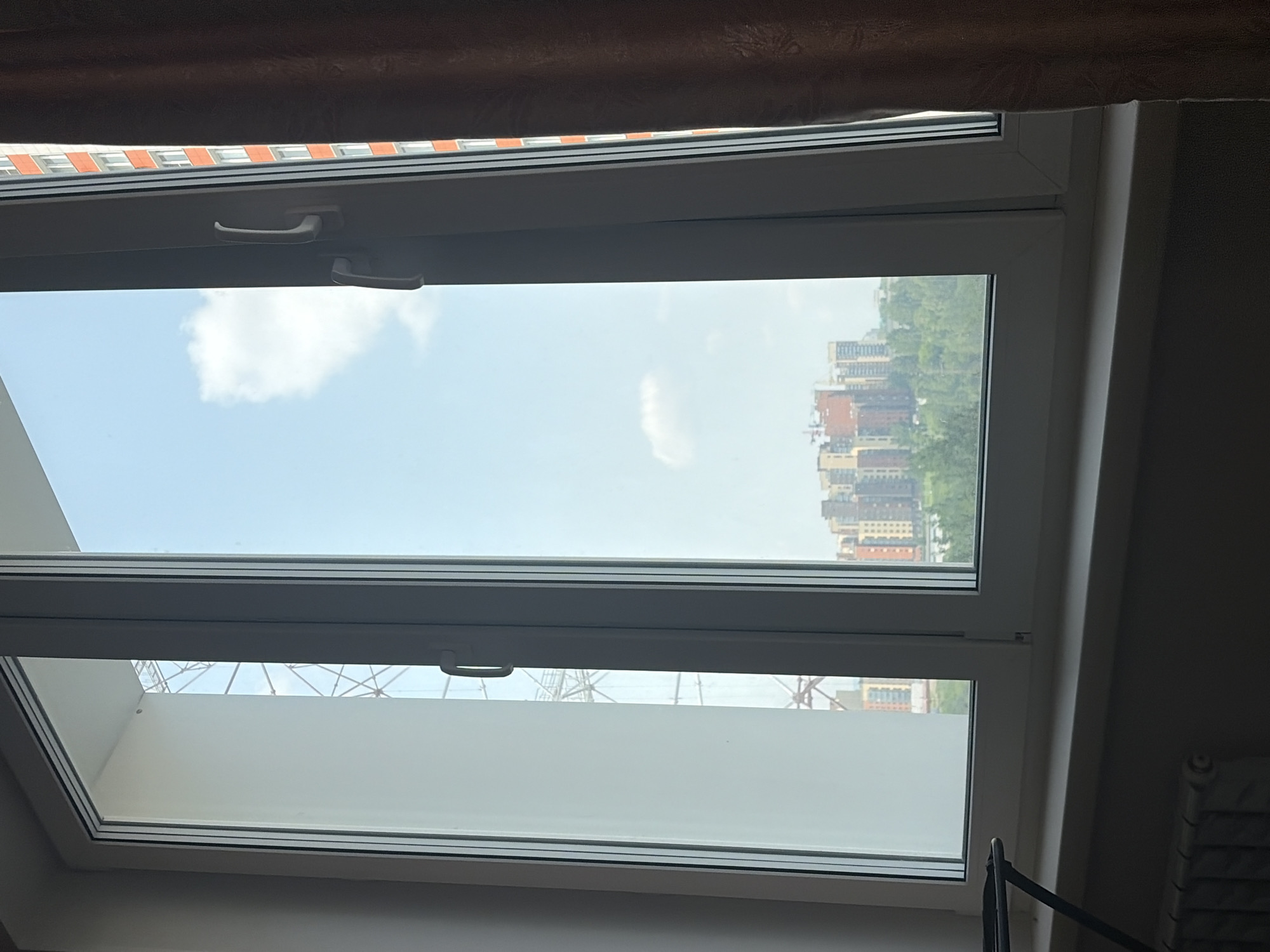 A view from a window shows a cityscape with multiple buildings under a bright sky with a few clouds. The window frame is white, and the left pane is slightly ajar. A brown curtain is partially visible on the right side of the frame. In the foreground, a radiator is visible below the window.