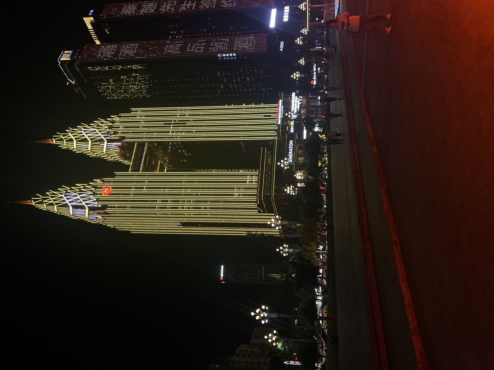 A nighttime exterior view captures a vibrant cityscape with prominent illuminated skyscrapers in Nan'an District, China. The central focus is a twin-peaked skyscraper adorned with a striking pattern of vertical golden lights, evoking a stylized flame or crown. Adjacent to it, another skyscraper glows with intricate patterns of red and white lights, forming a pixelated facade. To the left, a narrower, darker skyscraper stands, also displaying some illumination. In the foreground, a pedestrian area is visible, with several people walking and standing. Streetlights with multiple globes illuminate the scene, casting a warm glow on the trees and pavement. The street behind the pedestrian area shows traffic, with vehicles' headlights and taillights visible. The overall atmosphere is one of urban dynamism and modern architectural display under the night sky.