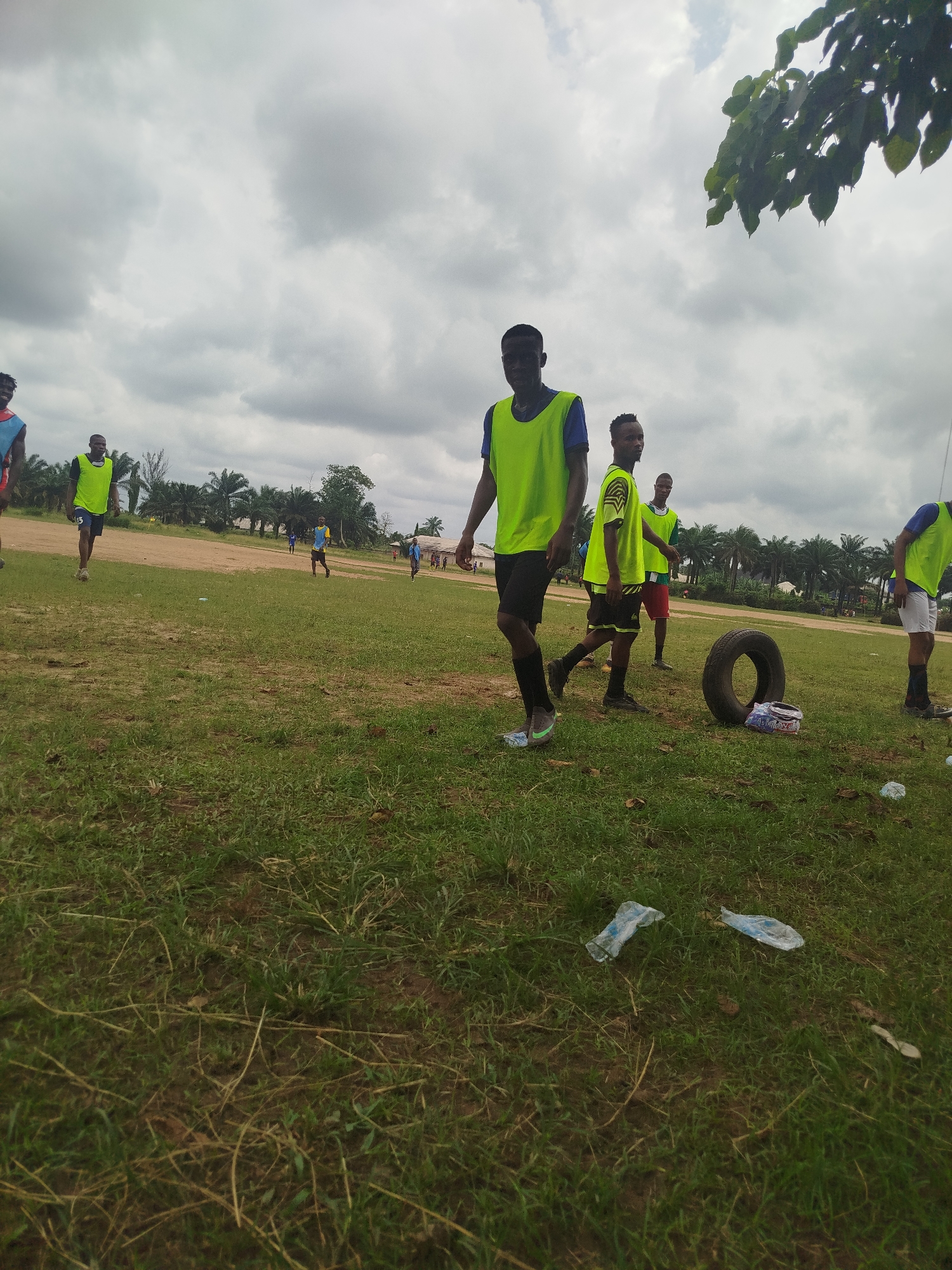 The image depicts a grassy field in Ikot Mbonde, Nigeria, under a cloudy sky. Several young men wearing neon-colored vests over blue shirts are scattered across the field. Some are walking while others appear to be engaging in light physical activity. In the midground, a black tire and a discarded snack wrapper rest on the grass. In the background, palm trees, a dirt road, and a building can be observed. Litter, including plastic bottles, is visible on the ground in the foreground.