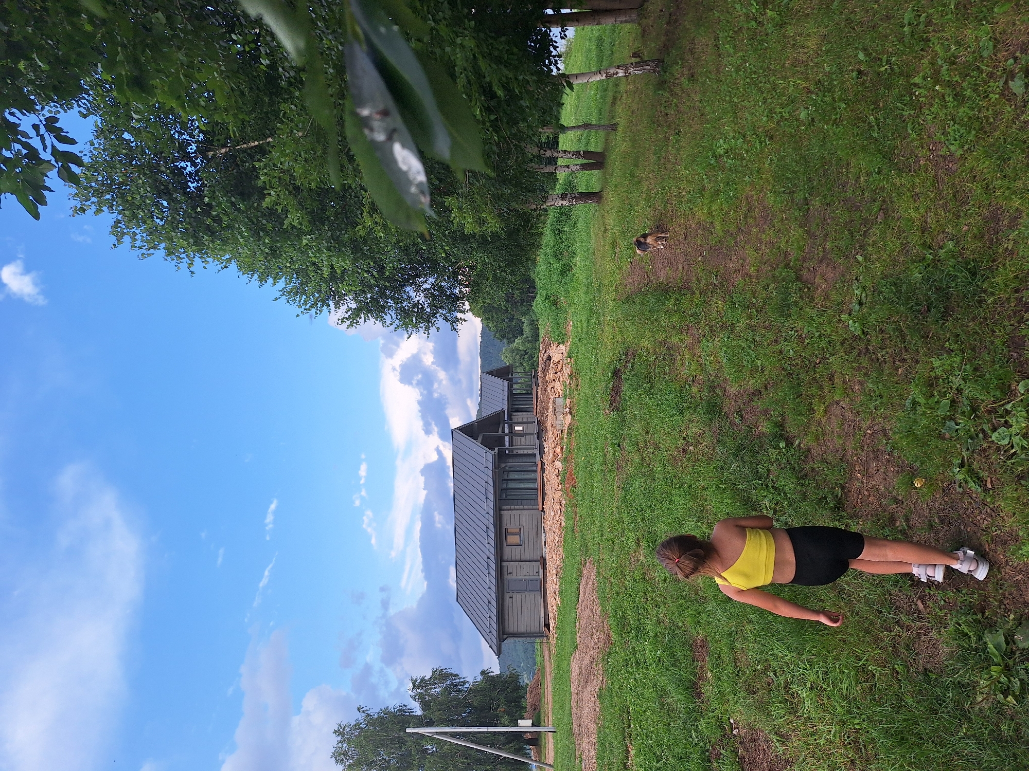 In Atamanovka, Russia, on a partially constructed property with green grass and dirt paths, a young girl with a ponytail, wearing a yellow one-shoulder top and black shorts, walks away from the camera. She is followed by a medium-sized dog with brown and black fur, walking on a dirt path. In the background, several modern, grey, single-story houses with dark roofs are visible. Tall green trees, including birches, line the right side of the frame. The sky is a mix of blue with scattered white clouds.