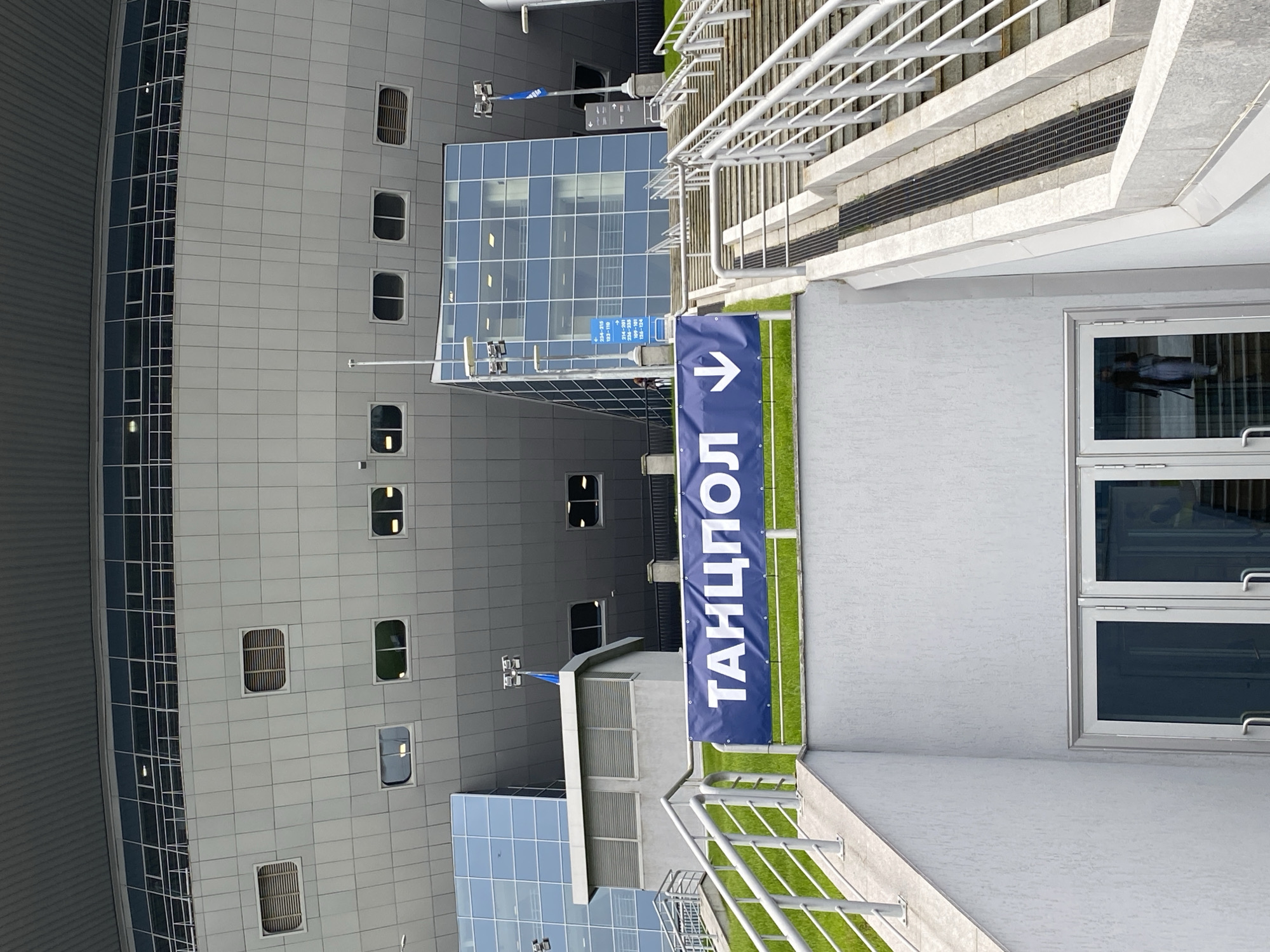 The image shows a view of the Pulkovo Airport in Saint Petersburg, Russia. A sign with blue background and white text "ТАНЦПОЛ" (dance floor) and a right-pointing arrow stands near a staircase with silver railings. The building's facade features a grid of windows and a glass-walled extension. There is also a small green area next to the staircase. A window on the right reflects a person and their belongings.