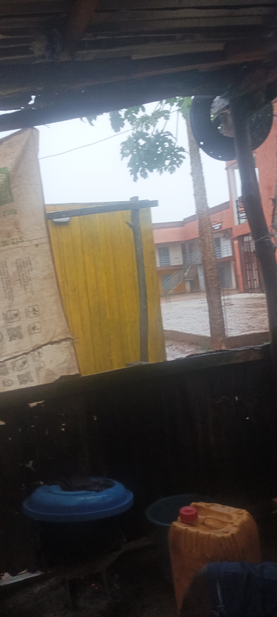 The image, taken from an interior location in Akwanga, Nigeria, shows an exterior view through a window. It's raining. Outside, a yellow corrugated structure stands next to a building with a brown and white exterior. A tree is also visible. Inside, in the foreground, are household items including a blue bucket and a yellow container.