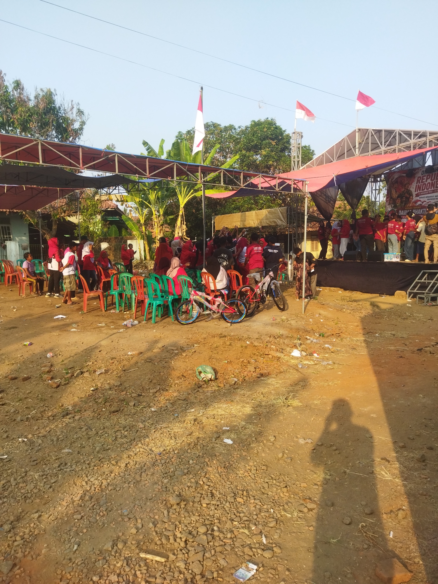 The image depicts an outdoor gathering under a temporary tent structure, likely in the city of Banjar, Indonesia, given the presence of Indonesian flags. A stage area is set up with a banner that reads "REPUBLIK INDONESIA" and "DARM BHAKTI." Several people are standing on the stage and in front of it, some appearing to be performers or speakers. In the foreground, a gathering of people are seated on plastic chairs. Two bicycles are parked near the seated individuals. The ground is unpaved and covered with dirt and small stones. The environment appears to be a community event, possibly a celebration or political rally, held during daylight hours, as indicated by the shadows cast by the people and the tent. The overall atmosphere seems to be one of public assembly.