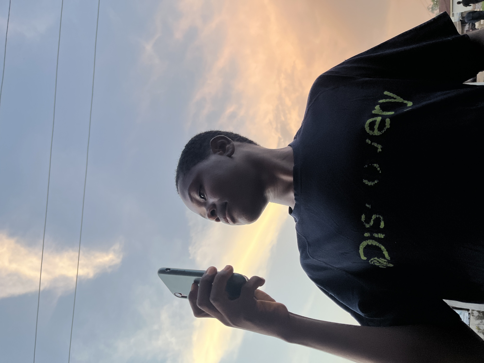 A low-angle, medium shot captures a young Black person from the chest up, holding a smartphone in their right hand and looking upwards and to the right. The subject is wearing a dark t-shirt with green lettering that reads "Discovery." The background consists of a dusky sky with soft clouds illuminated by the setting sun, casting a warm orange and yellow glow. Several power lines crisscross the sky. The scene appears to be outdoors, possibly in a residential area, with glimpses of rooftops and figures of other people in the far background on the right side of the frame. The image was taken in Gura Top, Nigeria.