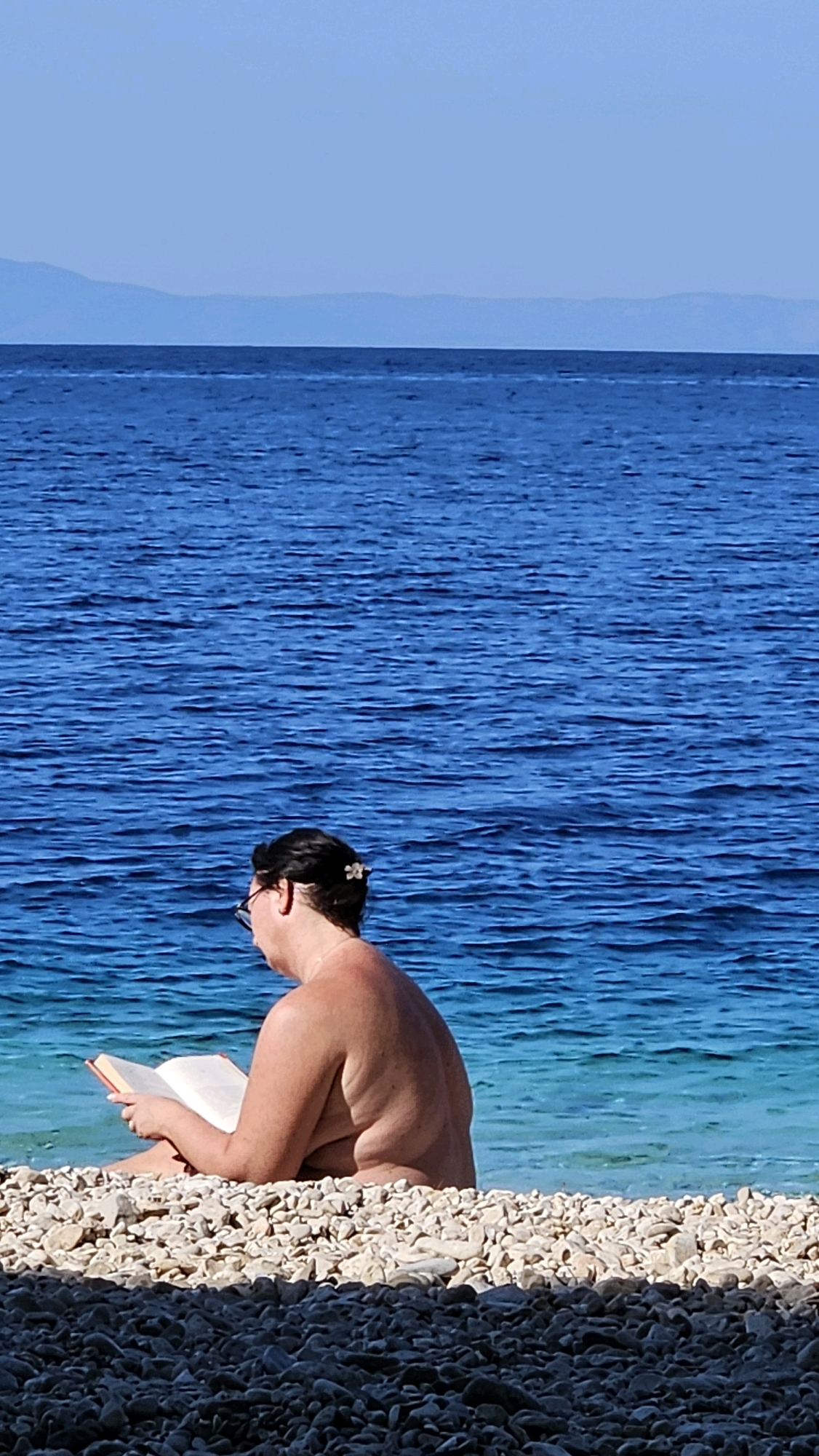 A person is sitting on a pebble beach in Općina Podgora, Croatia, reading a book. The person is facing to the left, with their back mostly to the camera. They are wearing glasses and have a flower in their hair. The sea is blue and calm behind them, with mountains visible in the distance on the horizon. The foreground of the image consists of a dark shadow cast over more pebbles.