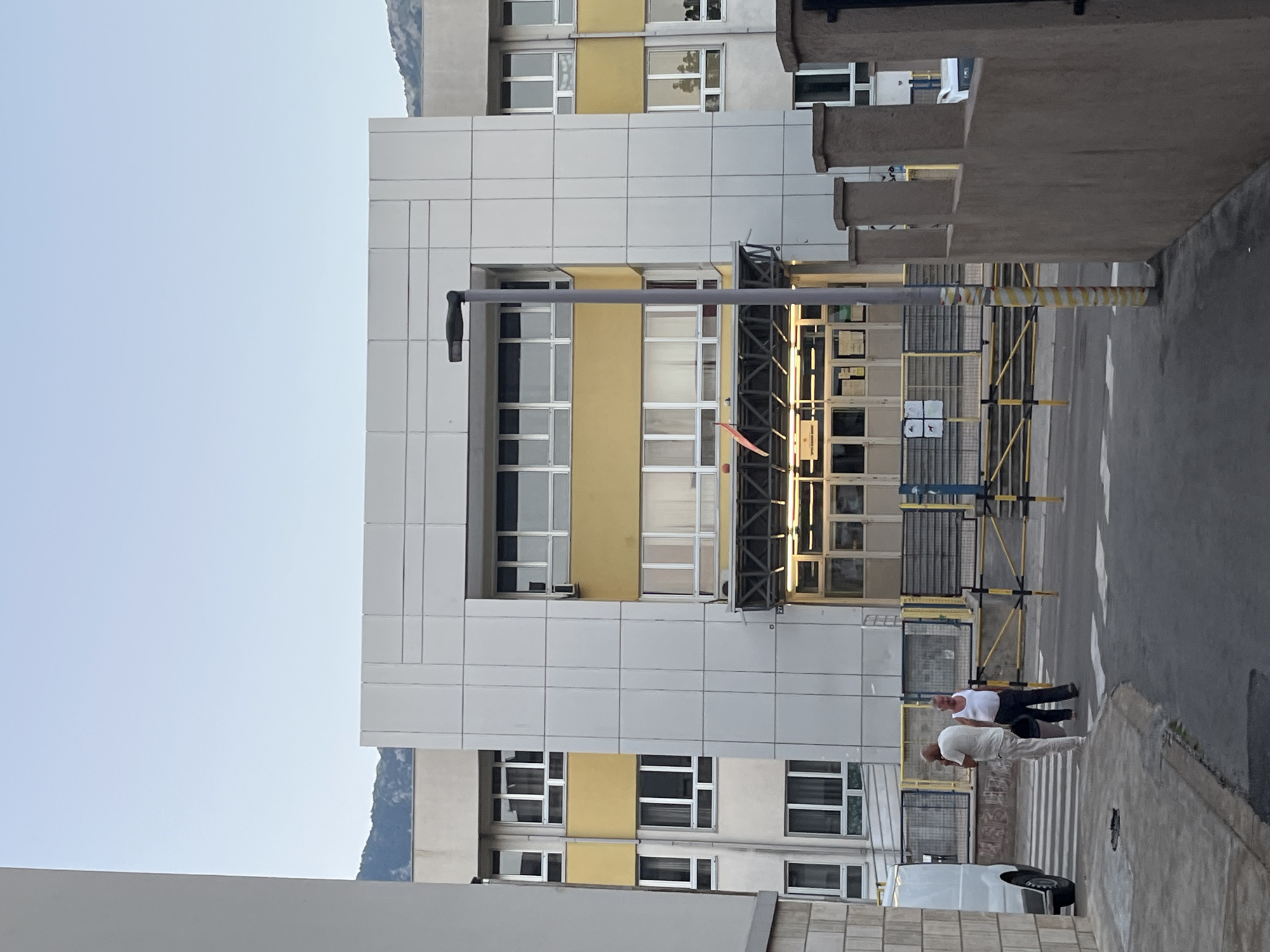The image shows the exterior of a building, likely a school, in Budva, Montenegro. The building is multi-story with a white and yellow facade, featuring numerous windows and a central entrance area with a canopy. There are two men standing on a crosswalk in front of the building, one in a white t-shirt and light pants, the other in a white tank top and dark pants. To the left of the building, a white van is parked. A street light stands in front of the building's entrance. A concrete wall with decorative pillars is visible in the foreground on the right. Mountains are visible in the background. The sky is clear and light blue.