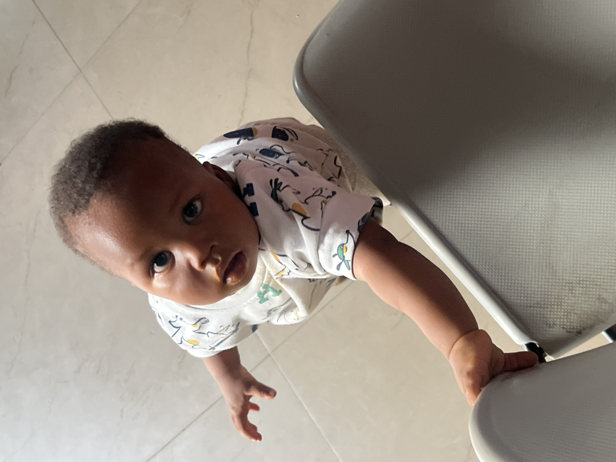A baby with dark hair and light brown skin, wearing a white shirt with a cartoon design, is positioned on a light-colored tile floor next to a white chair. The baby is looking up at the camera, gripping the edge of the chair with their right hand. The location is unknown.