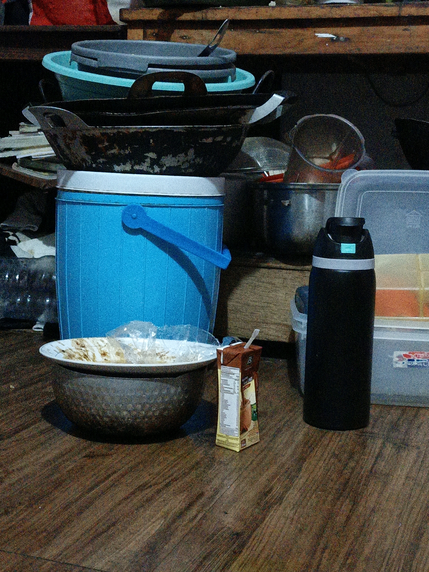 This image captures a cluttered, indoor scene that appears to be a kitchen or storage area. The foreground is dominated by a dark, textured bowl resting on a wooden floor, with a white plate holding some crumpled plastic and food remnants positioned on top of it. To the right of the bowl, a small, brown juice box with a straw sticks out. Behind the bowl and plate, a bright blue, ribbed bucket with a blue handle stands prominently. Further back, several cooking vessels, including a dark wok and a large grey basin, are stacked. In the background, there are more containers and what looks like shelves with various items. A black thermos with a silver band is also visible, positioned near a clear plastic container filled with what appear to be food items. The lighting is dim, casting shadows and creating a somewhat grainy texture across the image. The overall impression is one of a functional, possibly used, space. There are no people visible in the image. The presence of cooking utensils, food packaging, and storage containers suggests a domestic or commercial kitchen environment. The location is identified as West Jakarta, Indonesia.