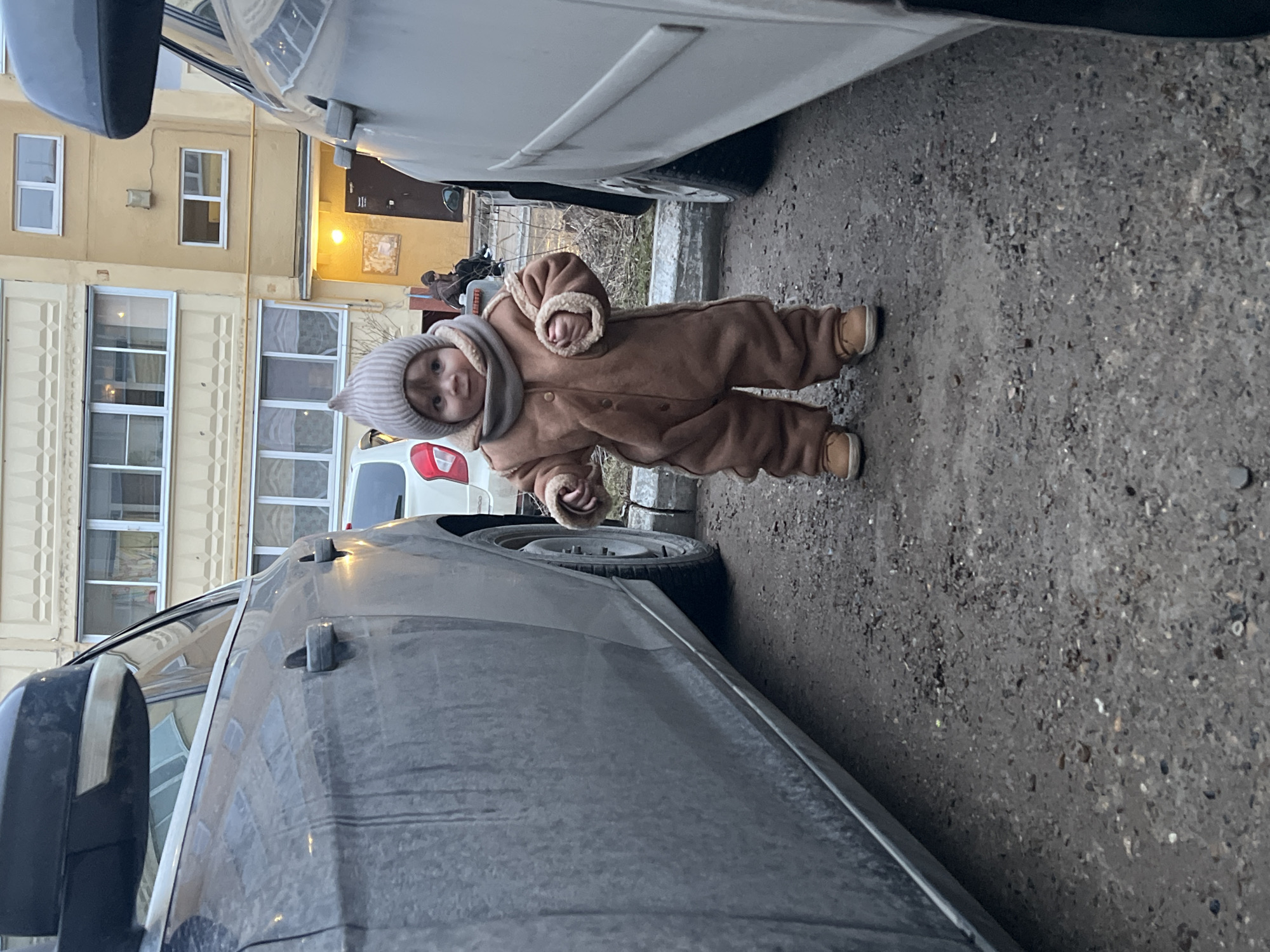 A toddler in a brown, furry-lined hooded snowsuit stands on a gray, textured asphalt surface between two parked vehicles. The vehicle to the right is light-colored, while the vehicle to the left is dark gray. The toddler faces the viewer, looking slightly to the left. In the background, a multi-story building with cream-colored facade and multiple windows is partially visible, suggesting a residential or commercial area. The photograph shows signs of urban environment like parking of the vehicles and architecture indicating that this photo was taken in a location with urban developments which align with the city location provided.