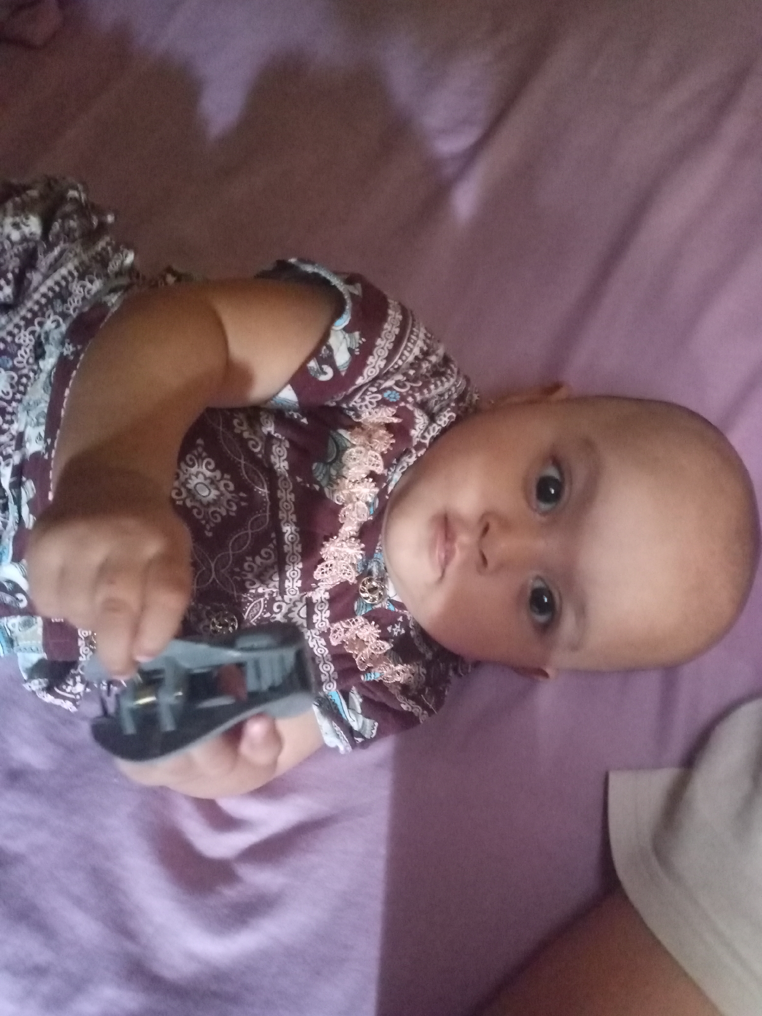 A baby is lying on a purple surface, possibly a bed or blanket. The baby is wearing a patterned shirt with maroon and white designs. The baby's head is turned to the left, and the baby is looking at the camera. The baby's right hand is holding a small, gray object, possibly a toy. The background is a solid purple color.