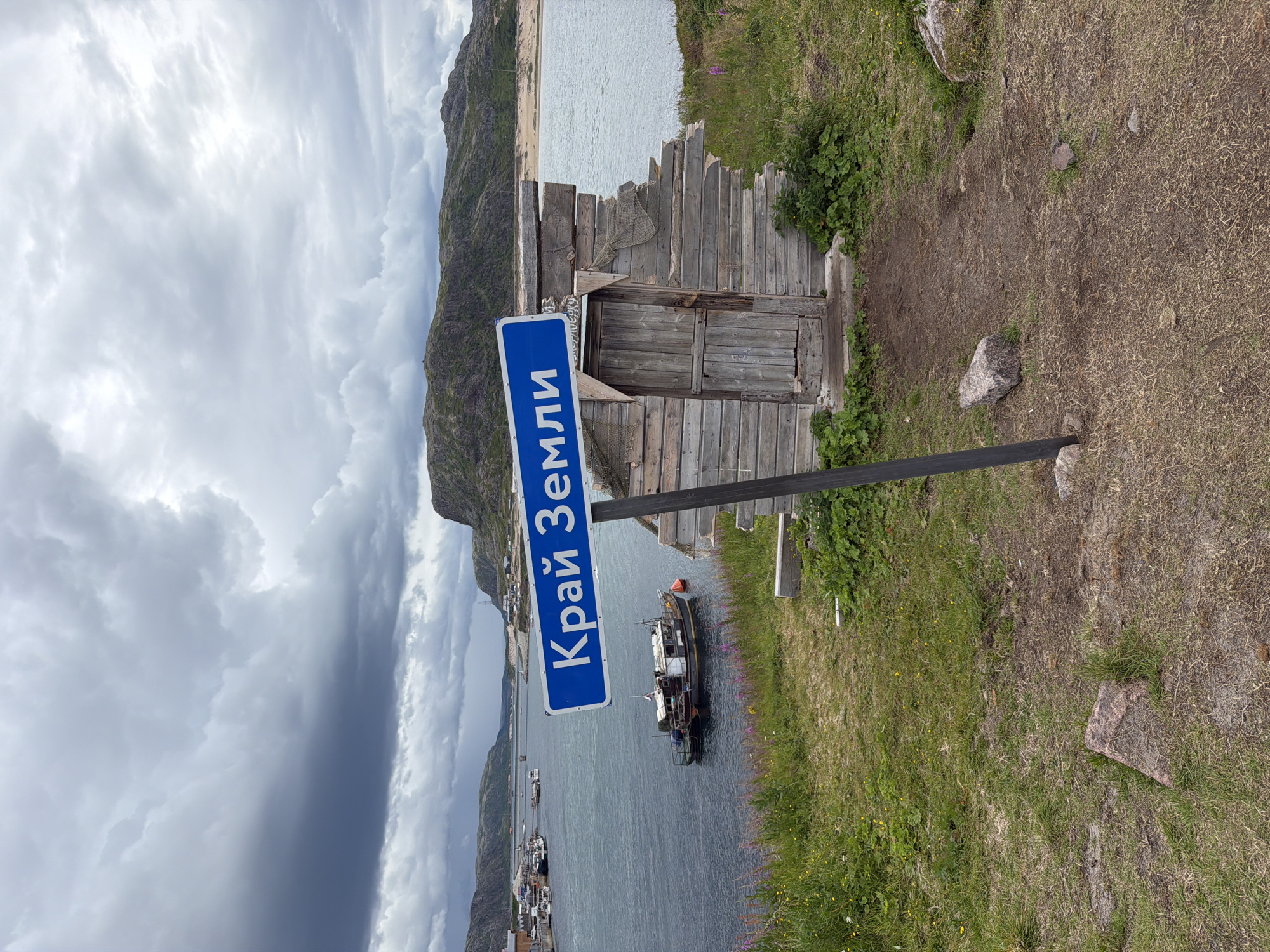 A weathered wooden shack with a broken roof stands on a grassy hill overlooking a body of water. A blue sign with white Cyrillic lettering, reading "Край Земли" (Edge of the Earth), is mounted on a dark pole in front of the shack. The water is dotted with several small boats, and in the distance, there are hills and a cloudy sky. The scene appears to be in Teriberka, Russia, a coastal town known for its remote location.