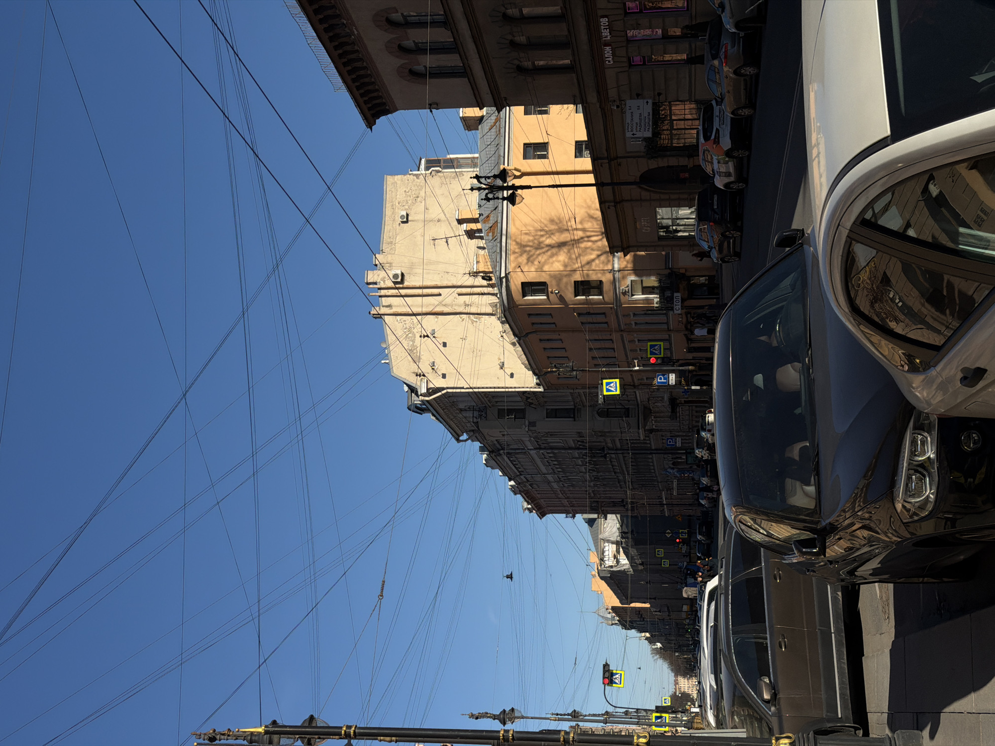 The image captures a street in Saint Petersburg, Russia, with buildings lining either side. Power lines crisscross the clear blue sky overhead. A row of parked cars occupies the right side of the street. Streetlights and traffic signals are visible, along with pedestrian crossing signs. The architecture of the buildings varies, with different heights and facade colors.