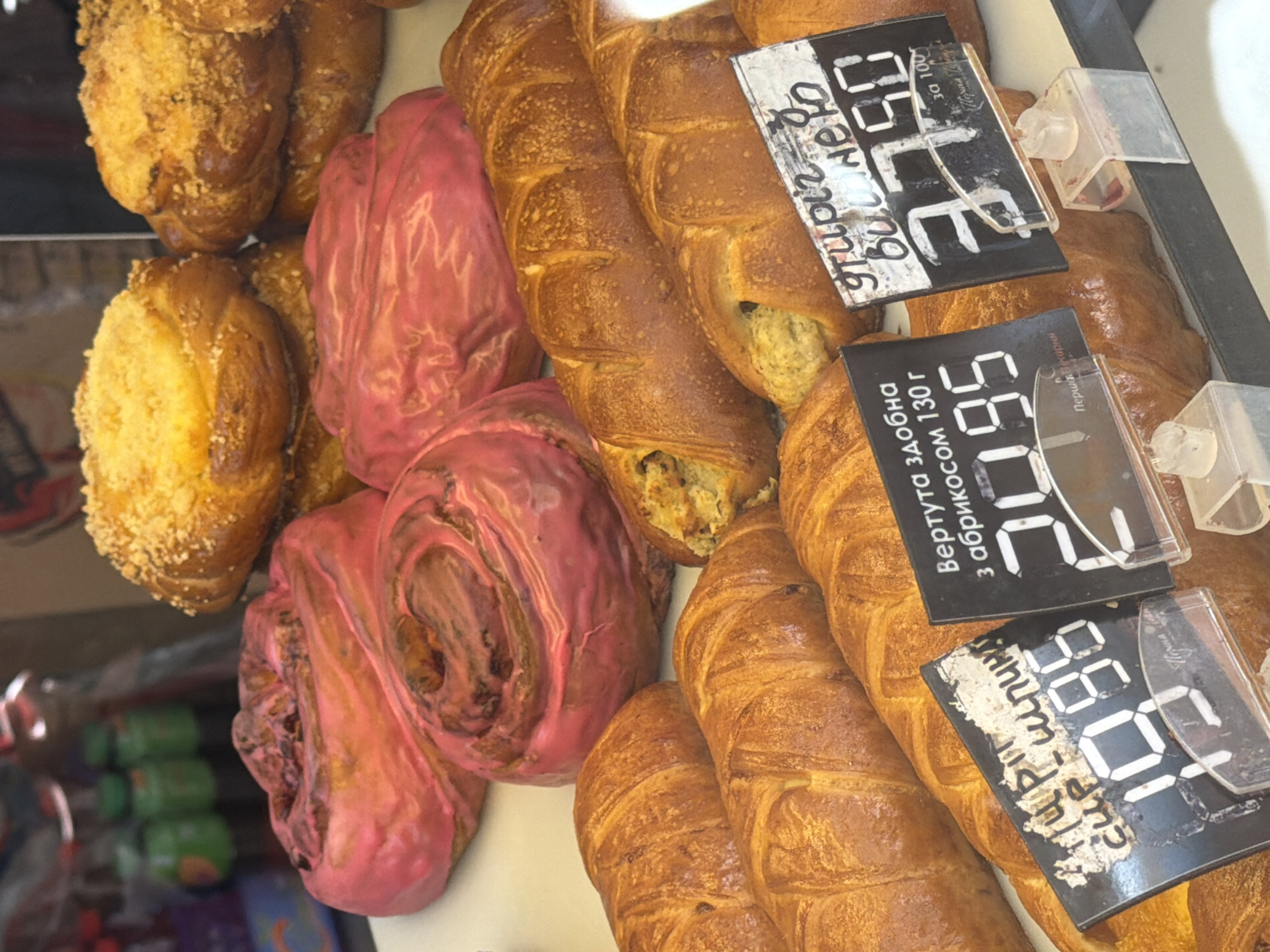 The image shows a display of baked goods, likely in a bakery or market, in the city of Stryi, Ukraine. In the foreground, there are several golden-brown croissants or similar pastries. Behind them, two long, braided pastries with visible filling are displayed. Further back, there are pink-frosted swirl pastries and some braided buns topped with crumbs. Two price tags are visible in the foreground. One tag has handwritten text and the number "19.95" displayed digitally. The other tag has handwritten text and the number "37.90" displayed digitally. The environment appears to be an indoor setting with a shelf or display case.
