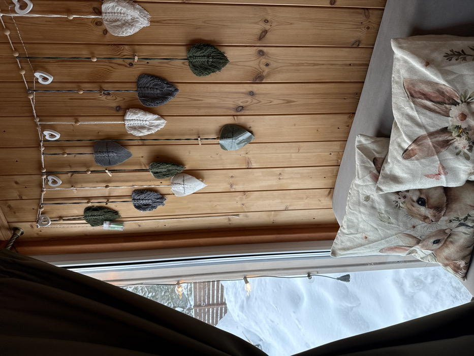 This image depicts an interior view within Sysert, Russia, looking out onto a snow-laden landscape. The right side of the frame features a natural wood plank wall, from which a decorative garland is suspended. This garland consists of a string with regularly spaced wooden beads and white heart-shaped ornaments, supporting twelve macrame leaf-shaped decorations in shades of dark green, gray, and off-white. Several leaves are secured with small wooden clothespins; one off-white macrame leaf exhibits the inscription "handmade with love," while a single dark green leaf below it appears to be fashioned from folded fabric. Below this, the edge of a gray sofa is visible, holding two light-colored pillows decorated with illustrations of rabbits and botanical elements. To the left, a window, partially framed by a dark brown curtain, reveals an exterior scene dominated by deep snow. A wooden fence is visible in the distance, partially buried, and subtle string lights are discernible through the windowpane.