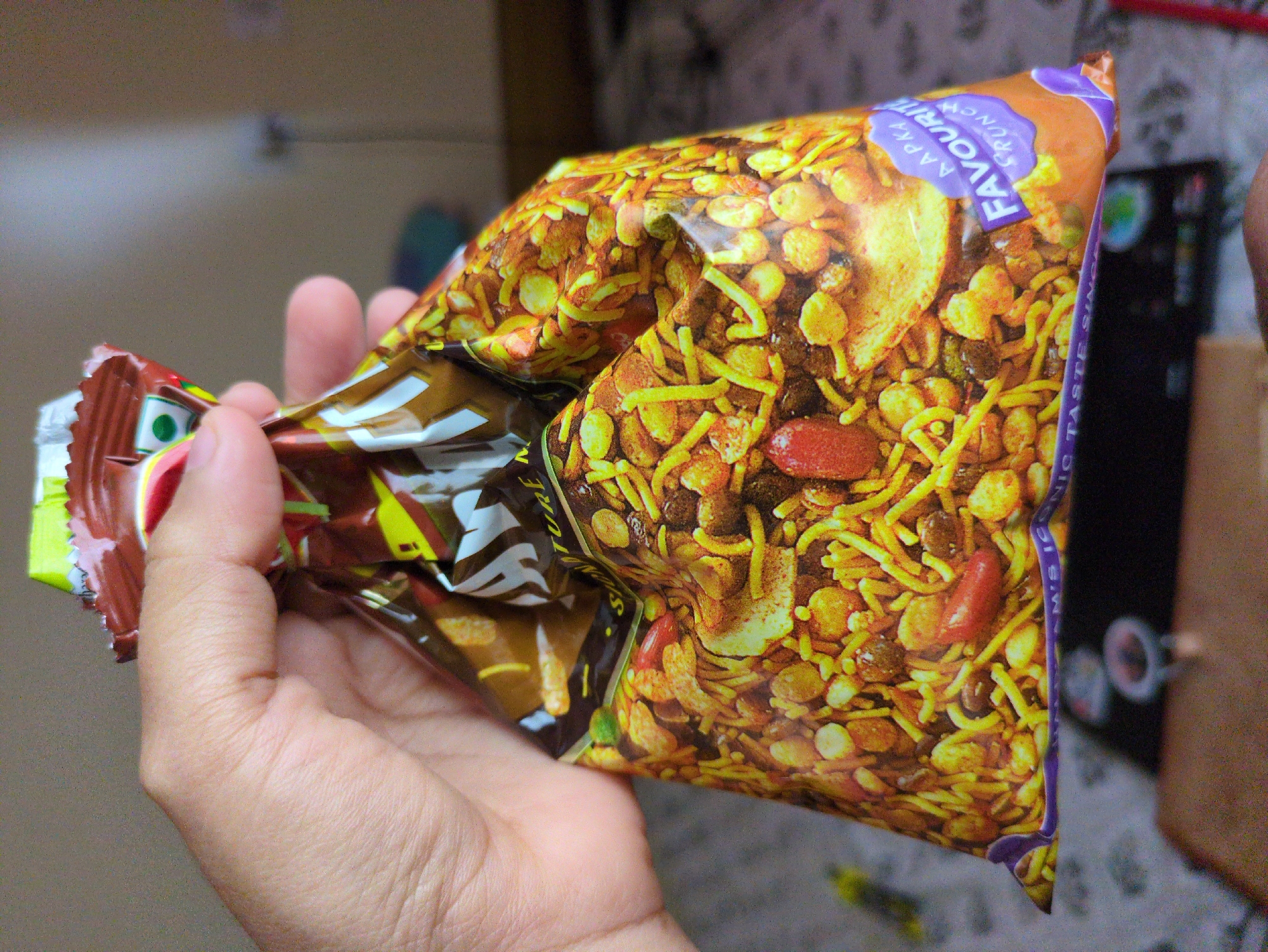 A hand holds a crushed bag of savory Indian snacks, likely Namkeen or Chivda, with a colorful, crumpled wrapper. The bag is partially open at the top, revealing a mixture of fried chickpea flour noodles, lentils, peanuts, and other crunchy ingredients. The wrapper features a brand name that is partially obscured but includes "ATURE" and a purple seal with "AAPKA FAVOURITE CRUNCH". The background is blurred, suggesting an indoor setting, with a textured surface and some indistinct objects, possibly furniture. The lighting is soft, with a slight warm cast. This scene is not specifically located in the city of Bhagsu, India; it is a general indoor scene.