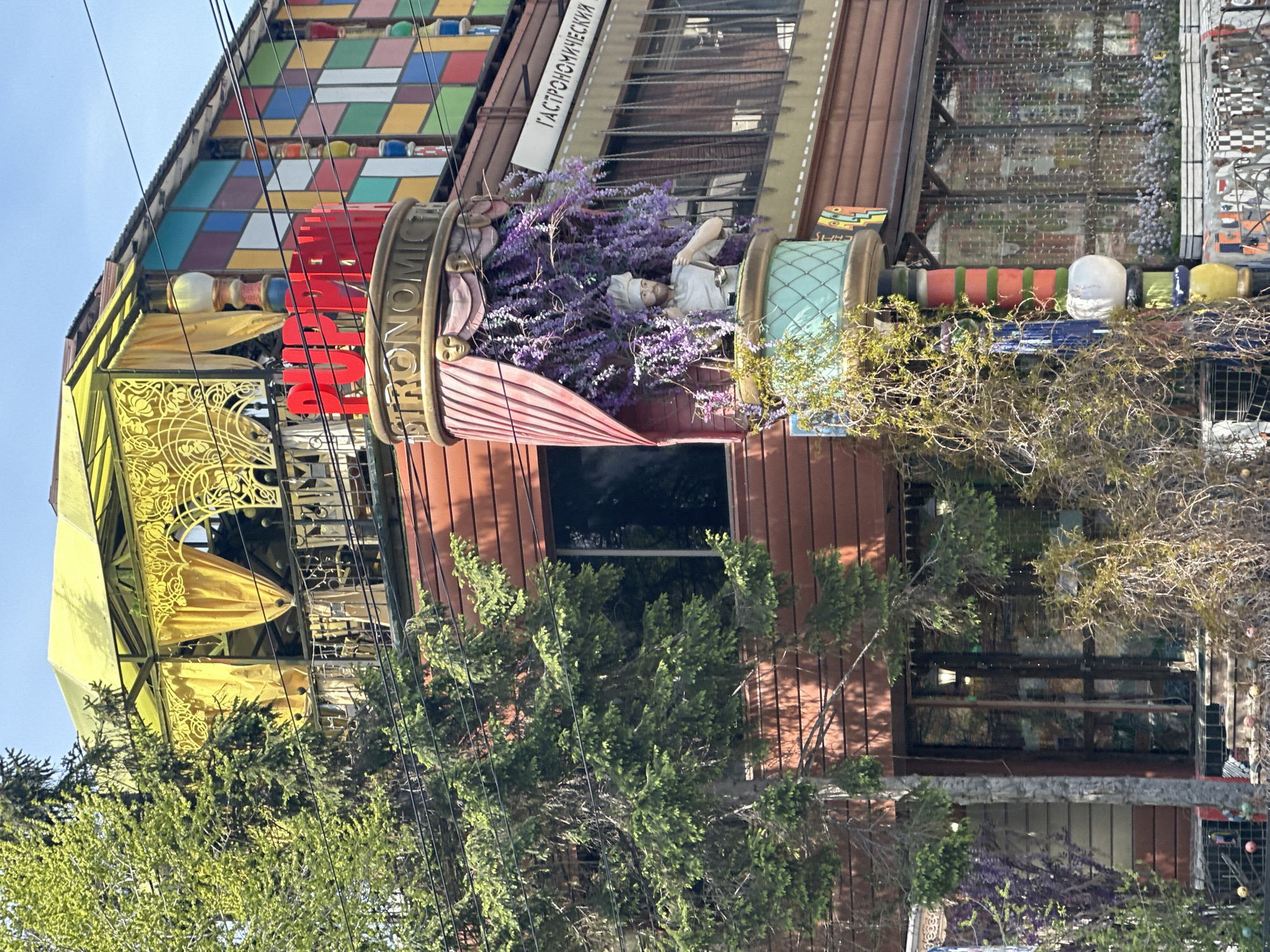 The image captures a building with distinctive and whimsical architectural features in Novosibirsk, Russia. The building displays a multi-colored patchwork facade, complemented by a yellow awning with ornate detailing. A sizable, cylindrical structure with the word "GASTRONOMIC" vertically aligned is prominently visible. In this structure, there is a figure of a chef surrounded by purple flowers. Trees partially obscure the lower portion of the building, and overhead power lines are visible in the foreground. The building's design suggests it is a commercial establishment, possibly a restaurant or culinary-themed business.