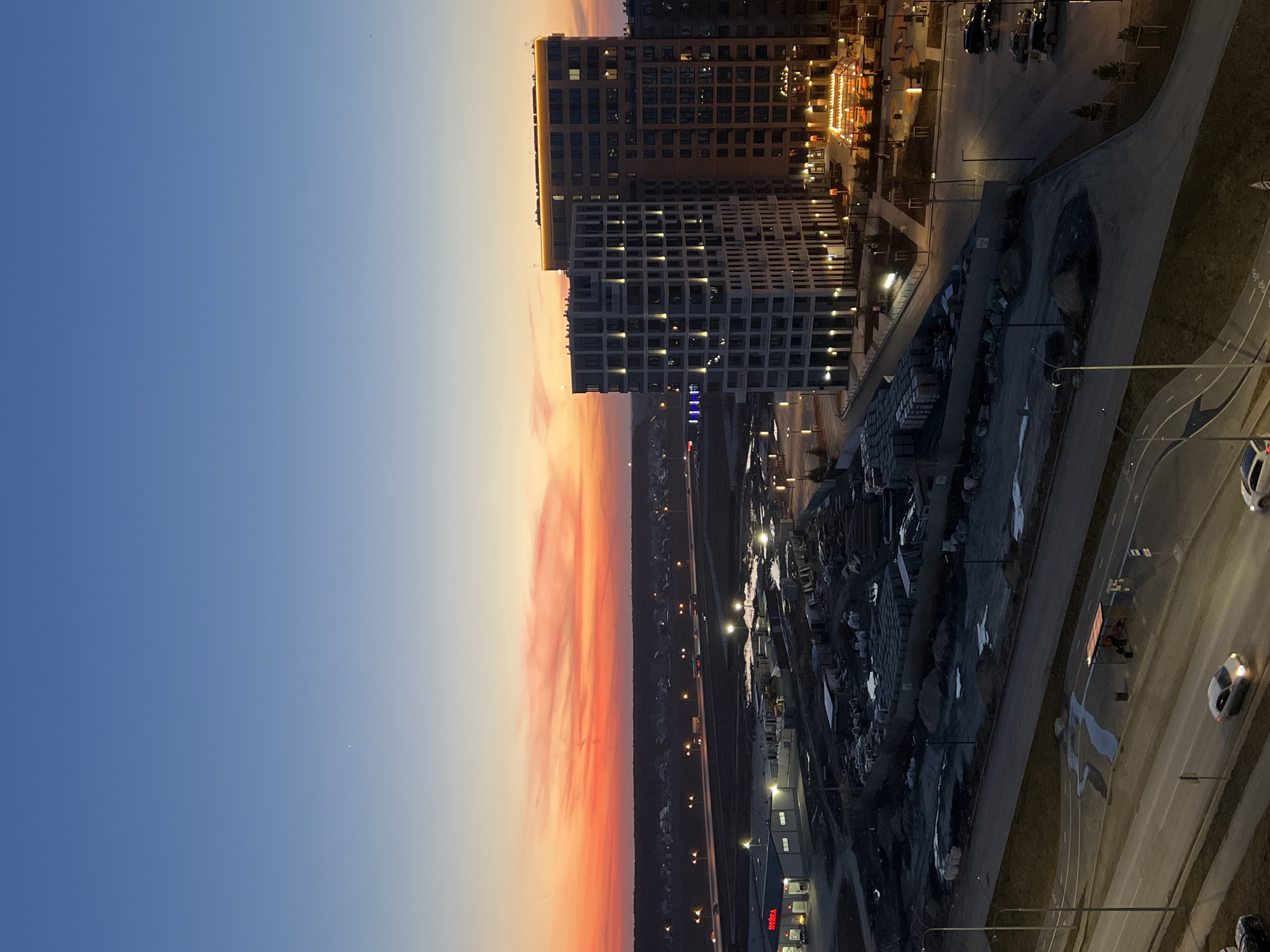 The image depicts a cityscape during a sunset in Tyumen, Russia. On the right side, two multi-story buildings are visible, illuminated with internal lights. Adjacent to the buildings is a parking lot with several vehicles, streetlights, and landscaping. Below the parking lot, a road is shown with cars and markings. On the left side of the image, the sky transitions from a deep blue to a fiery orange and red gradient, indicating sunset. A highway with occasional lights and distant buildings can be seen below the sunset sky.