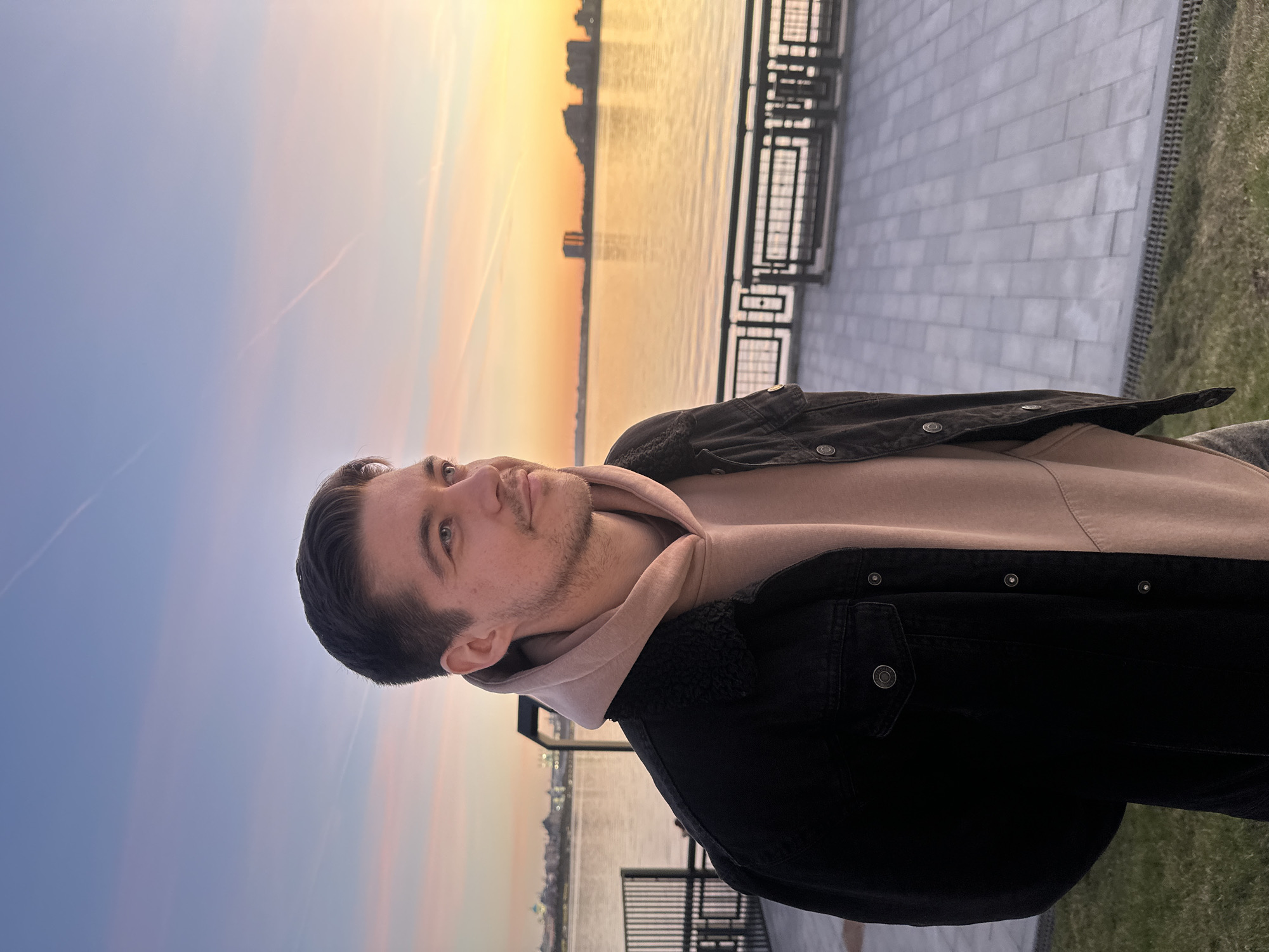 The image features a young man captured outdoors on what appears to be a waterfront promenade, with the scene set during either sunrise or sunset. The man, positioned on the left, has short dark hair and light facial hair, looking slightly upwards and to his right with a contemplative or observant expression. He is dressed in a light beige hooded sweatshirt layered under a dark, possibly black or charcoal grey, denim-style jacket that features a visible textured lining, likely shearling or fleece, around the collar.
The background is dominated by a large body of water, possibly a river or lake, which reflects the vibrant colors of the sky. Across the water, a distant city skyline with numerous buildings is silhouetted against the bright horizon. The sky exhibits a striking gradient, transitioning from a soft blue on the left to warm oranges, yellows, and pinks on the right, characteristic of twilight hours. Faint contrails are visible against the expansive sky.
To the man's right, a paved walkway, constructed from light-colored rectangular blocks, extends into the distance. This promenade is lined by a dark metal railing or fence that separates it from the water. A narrow strip of green grass is visible at the bottom right corner of the frame. The overall ambiance is tranquil and scenic, set in a waterfront area in Kazan, Russia. There is no visible text in the image.