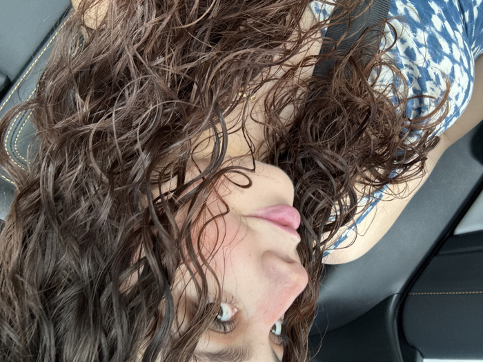 A woman with damp, dark curly hair is angled upside down, possibly in a car. She's wearing a blue and white patterned top and a seatbelt. Her face is visible, showing her eyes, nose, and lips. The background includes a black car interior, suggesting a relaxed or candid moment.