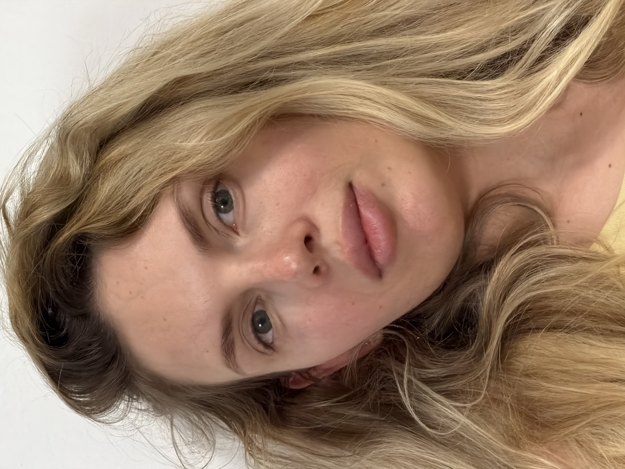 This media file presents a close-up, horizontally oriented portrait of a person, likely female, with long, wavy blonde hair. The subject looks directly at the camera with a neutral, relaxed expression. Her head is tilted, with her face occupying the central and lower-left portion of the frame. She has fair skin, adorned with light freckles, and natural-looking lips that are slightly parted. Her eyes are a soft green or grey-blue color.
Her voluminous blonde hair, which shows variations in shade from lighter ends to slightly darker roots, cascades around her face and spreads across the frame, filling much of the background. The detailed crops highlight the natural texture and slight waviness of her hair, as well as the fine details of her skin and features. A glimpse of light yellow fabric, possibly clothing or a pillow, is visible on the right side of the image, near her neck. The background behind her hair and face is plain white, suggesting an indoor setting. The lighting is soft and even, creating a gentle and intimate atmosphere. No text is visible in the image. The depicted scene is set in Budva, Montenegro.